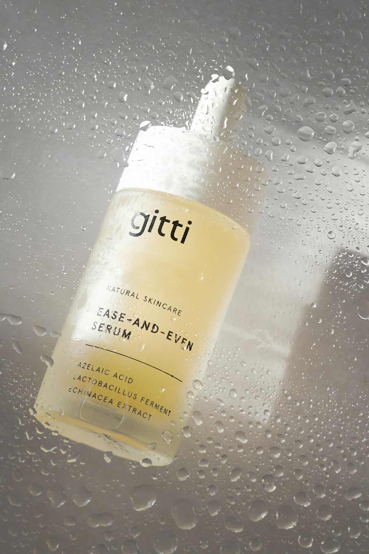 Ein Flakon des Ease-and-Even-Serums steht hinter einem nassen Glas mit Wassertropfen und präsentiert sein Etikett und seine Formel, die die Talgproduktion ausgleichen und Hautunreinheiten reduzieren soll.