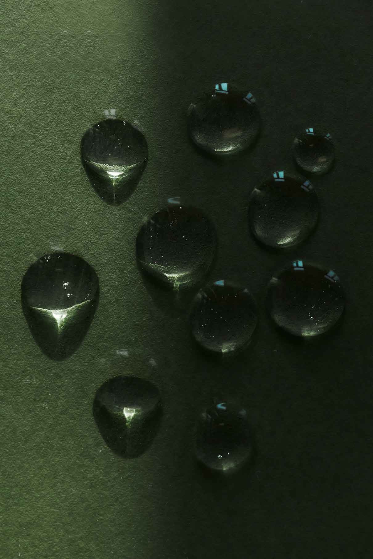 Nahaufnahme von Tautropfen auf einer tiefgrünen, strukturierten Oberfläche, deren Feuchtigkeit durch einen vertikalen Schatten hervorgehoben wird - ein Hinweis auf die erfrischende Feuchtigkeit des Glow-and-Protect-Serums.