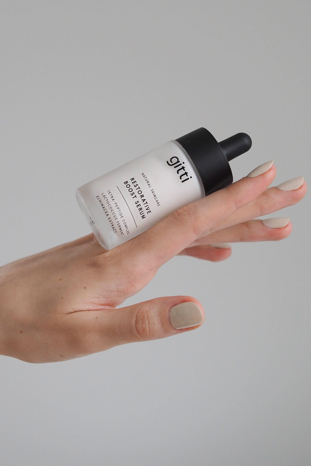 Eine Hand mit ordentlich manikürten Nägeln balanciert eine Flasche Restorative Boost Serum mit schwarzem Tropfverschluss vor einem schlichten hellgrauen Hintergrund, der Pflege für Feuchtigkeit und Elastizität der Haut hervorhebt.