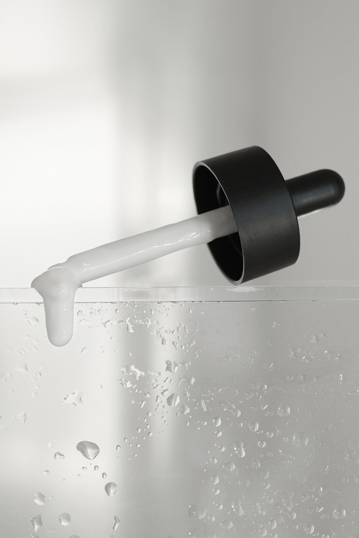 Ein schwarzer Tropfer mit weißer Flüssigkeit aus dem Restorative Boost Serum, das die Hautfeuchtigkeit und -elastizität erhöhen soll, ruht auf dem Rand einer klaren, feuchten Oberfläche.