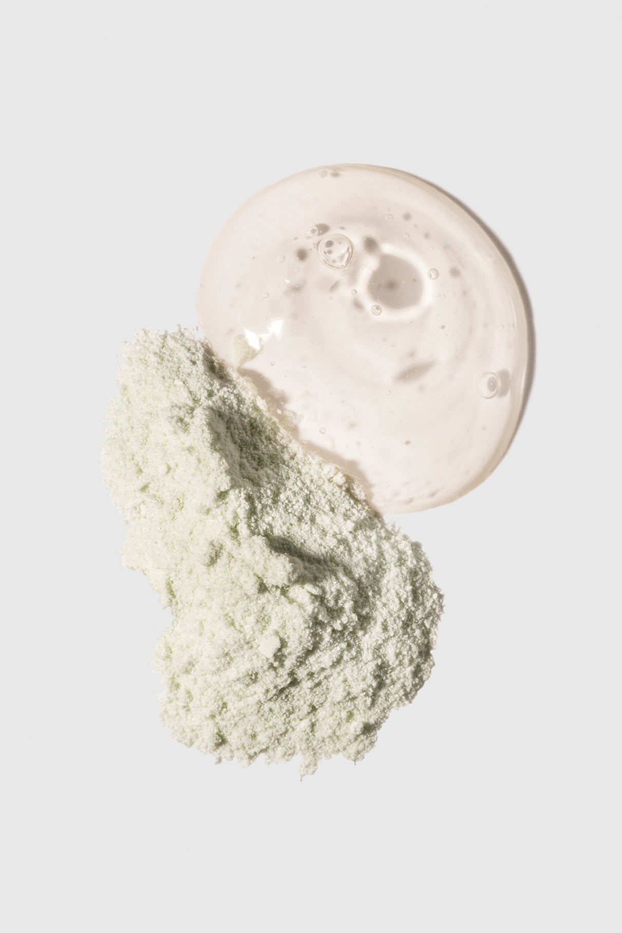 Ein blassgrüner Ultimate Glow Duo Puder sitzt neben einer Pfütze mit klarem Serum auf einem schlichten weißen Hintergrund und hebt moderne Hautpflege-Essentials hervor.