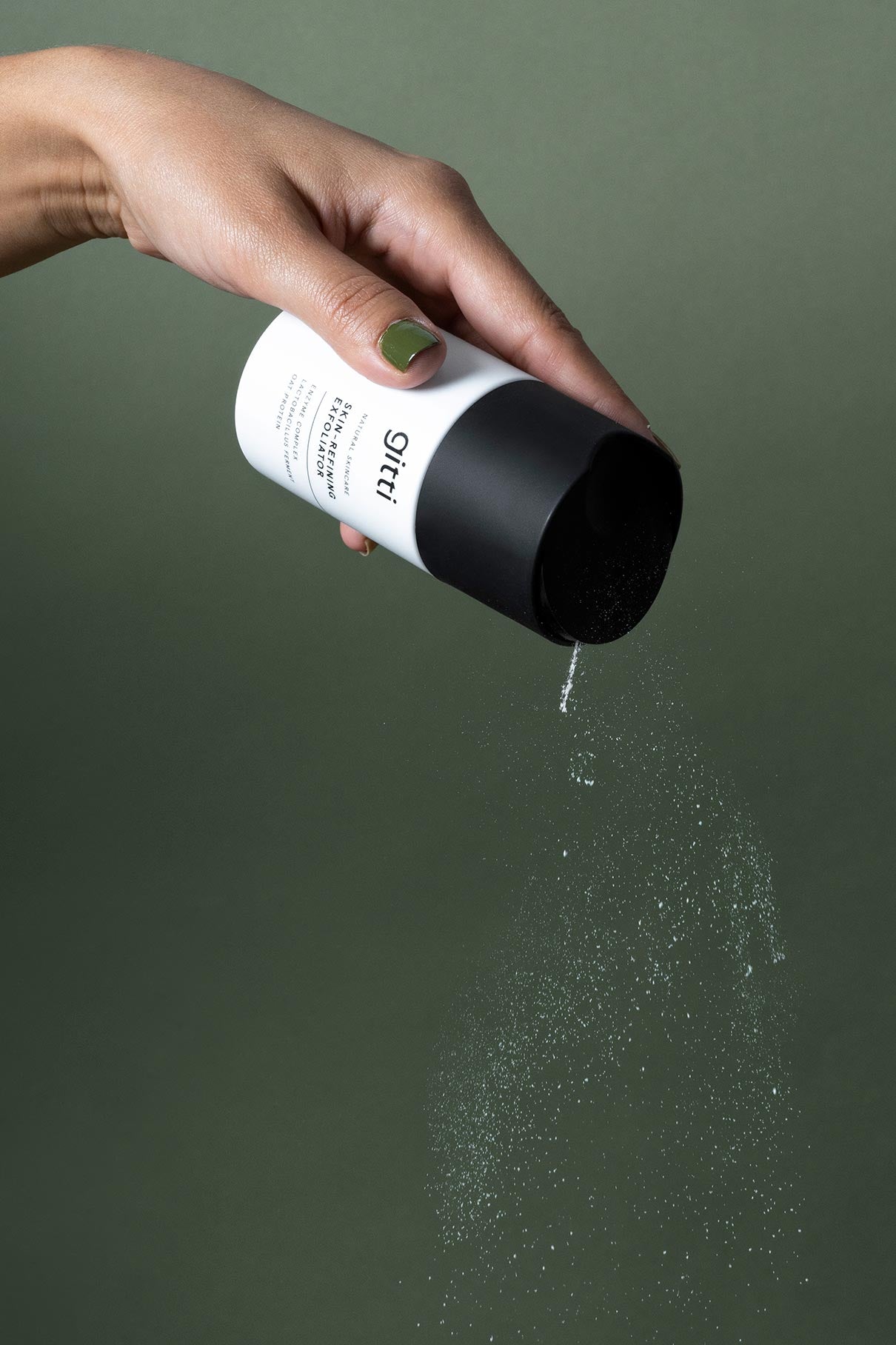 Eine Hand kippt einen schwarz-weißen Behälter und streut hautverfeinerndes Peeling-Pulver vor einem schlichten grünen Hintergrund.