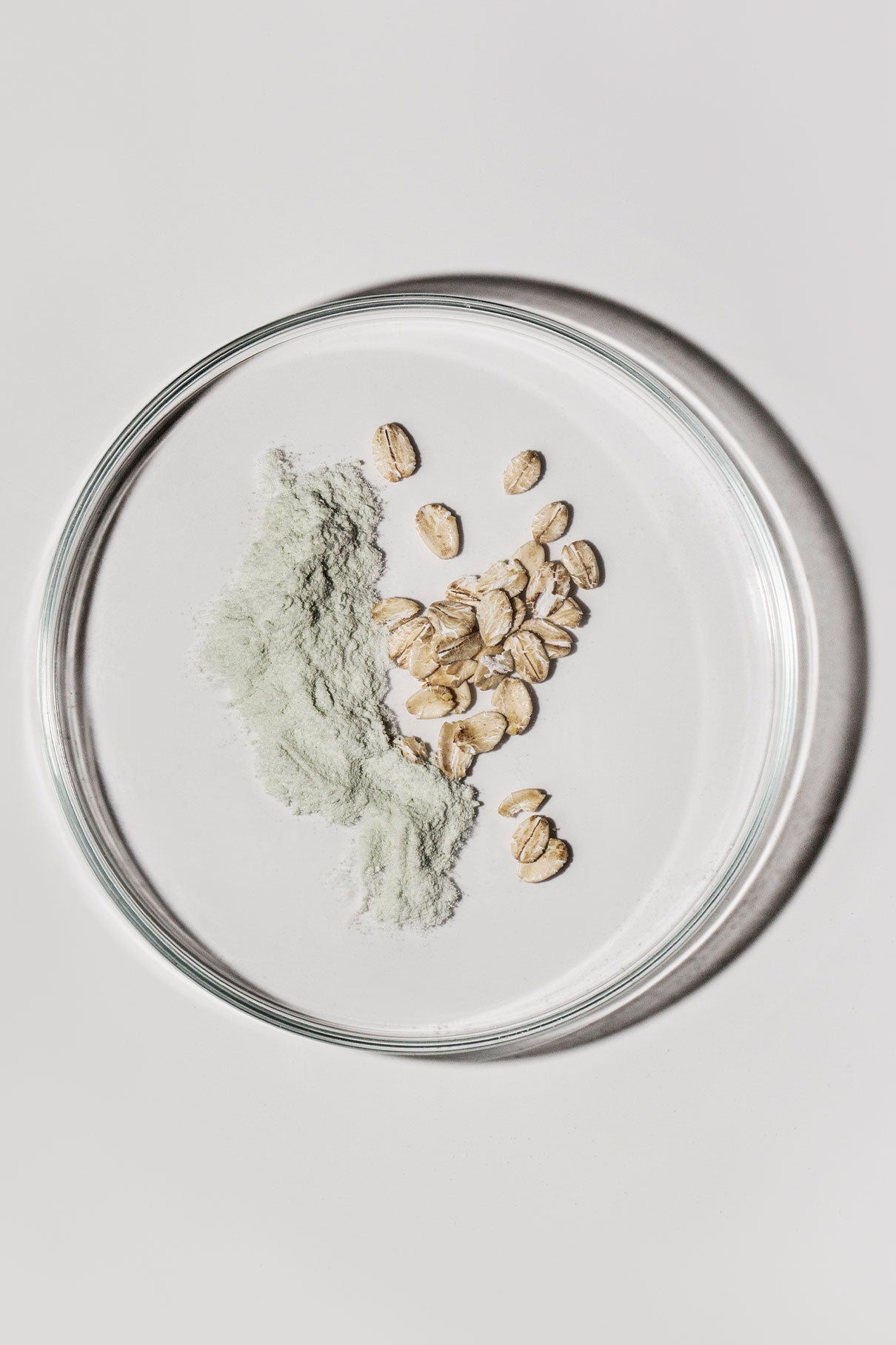 Eine gläserne Petrischale mit einem kleinen Haufen grünem Skin-Refining Exfoliator-Pulver und ganzem Hafer auf weißem Grund - ideal für die sanfte Reinigung als natürliche Peeling-Maske.