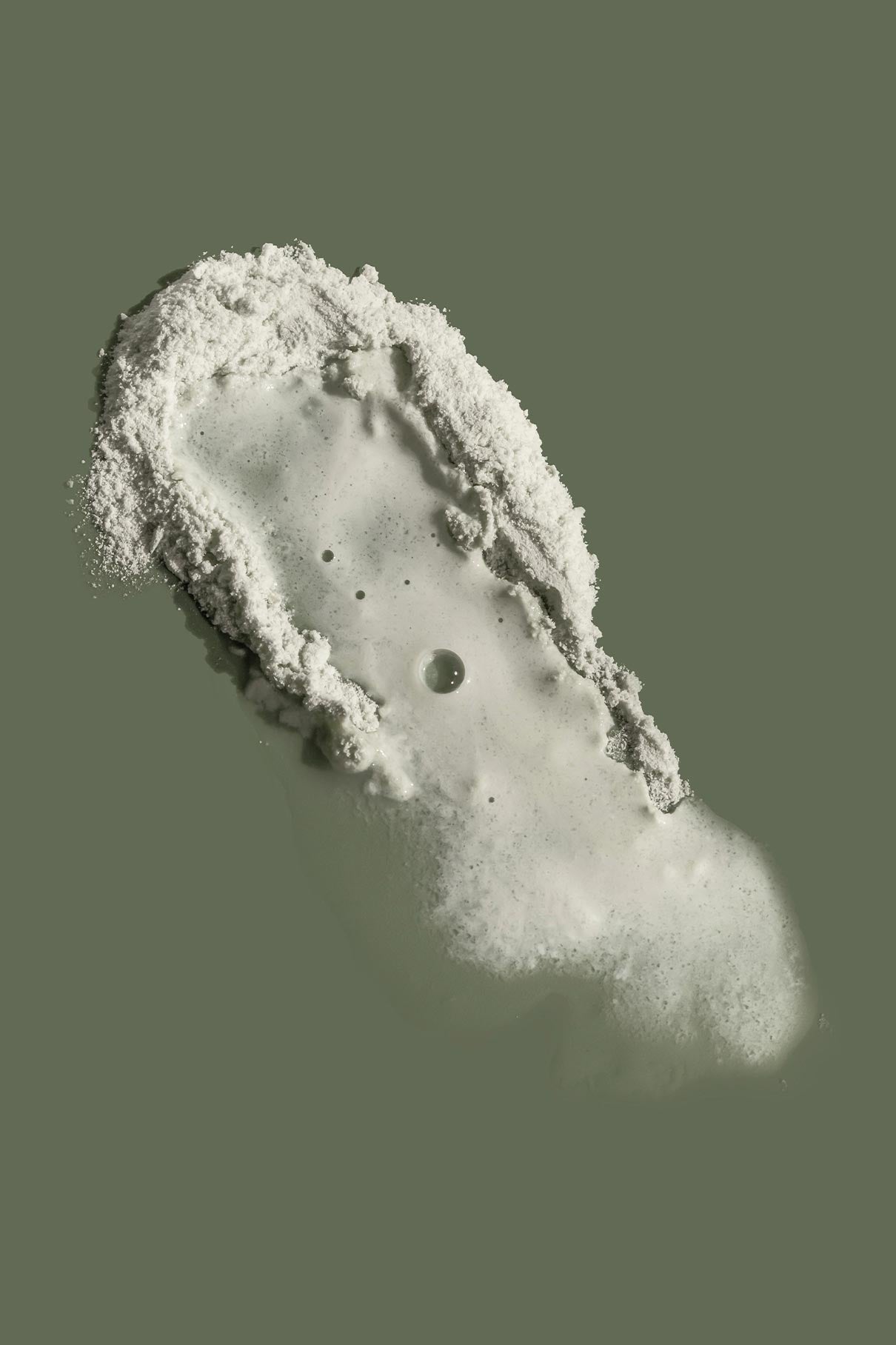 Ein fußabdruckförmiger Fleck aus weißem Pulver und Flüssigkeit auf grünlichem Grund erinnert an die sanfte Wirkung des Skin-Refining Exfoliator, perfekt für die sanfte Reinigung oder als Peeling-Maske.