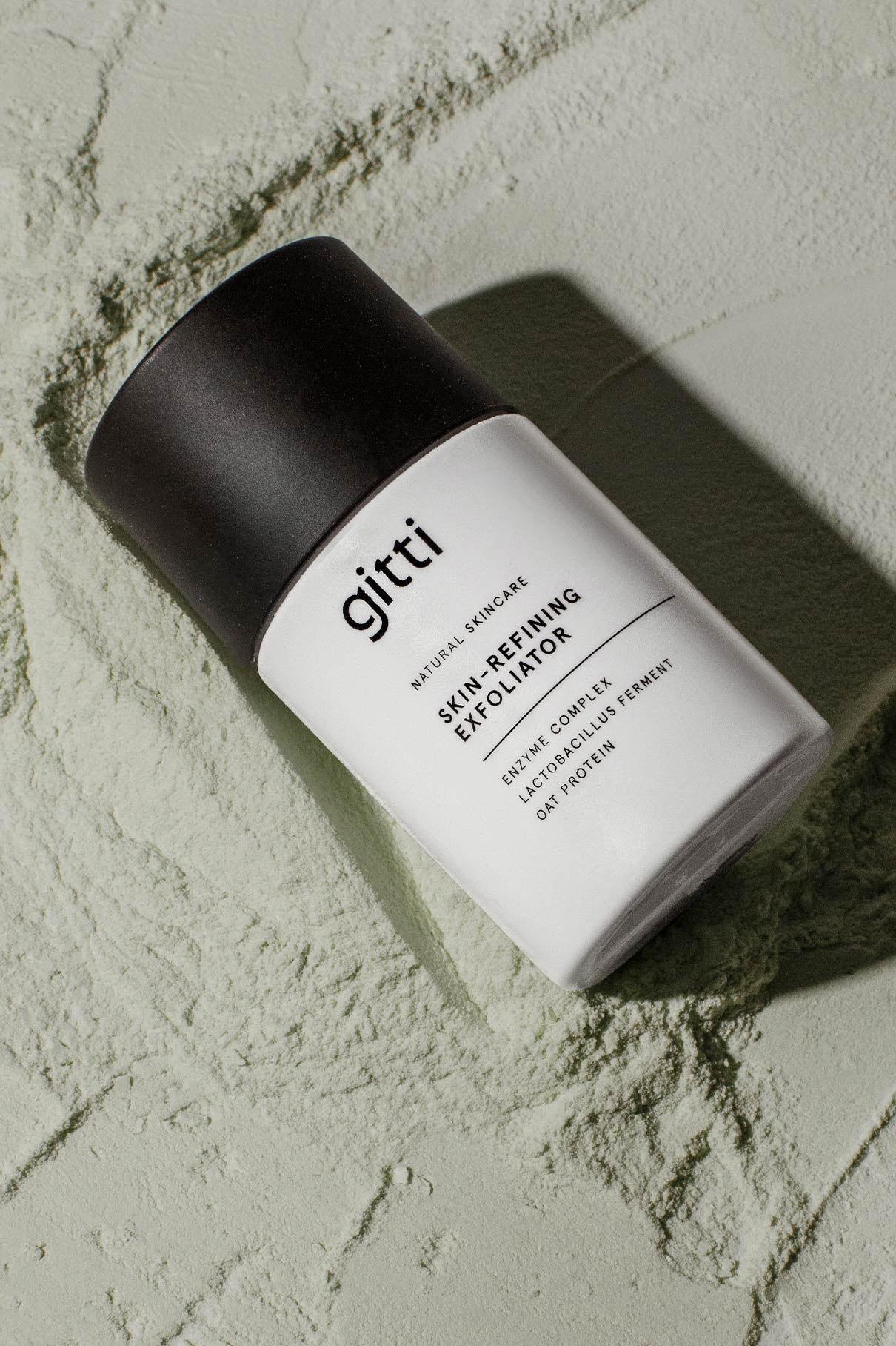 Ein weißes Fläschchen mit der Aufschrift "Skin-Refining Exfoliator" und einer schwarzen Kappe ruht auf einem strukturierten, hellgrünen Pulver - ideal für die sanfte Reinigung und die Verwendung als Enzym-Peeling oder Peeling-Maske.