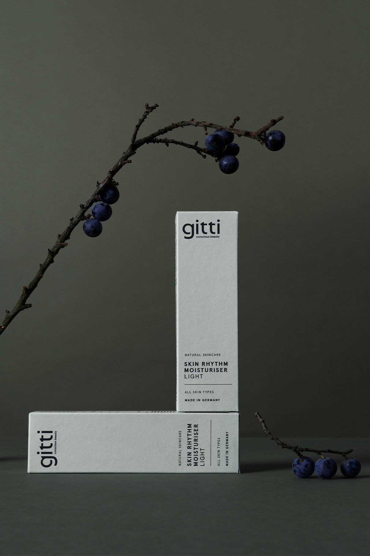 Zwei minimalistische Essential Routine Pflegeboxen von gitti stehen auf einer dunklen Fläche neben einem beerenreichen Zweig - perfekte Essentials für Ihre tägliche Beauty-Routine.