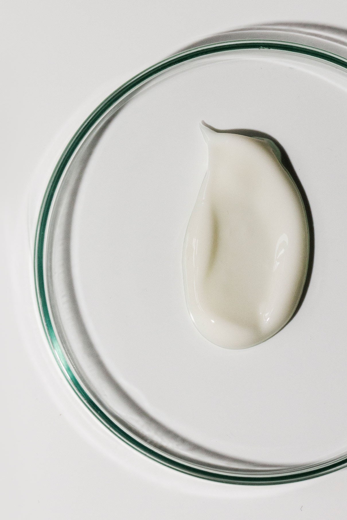 Skin Rhythm Moisturiser Light, eine Petrischale aus klarem Glas mit einer kleinen Menge weißer Creme zur Steigerung der Feuchtigkeit und Förderung eines gesunden Aussehens, ruht auf einer weißen Oberfläche.