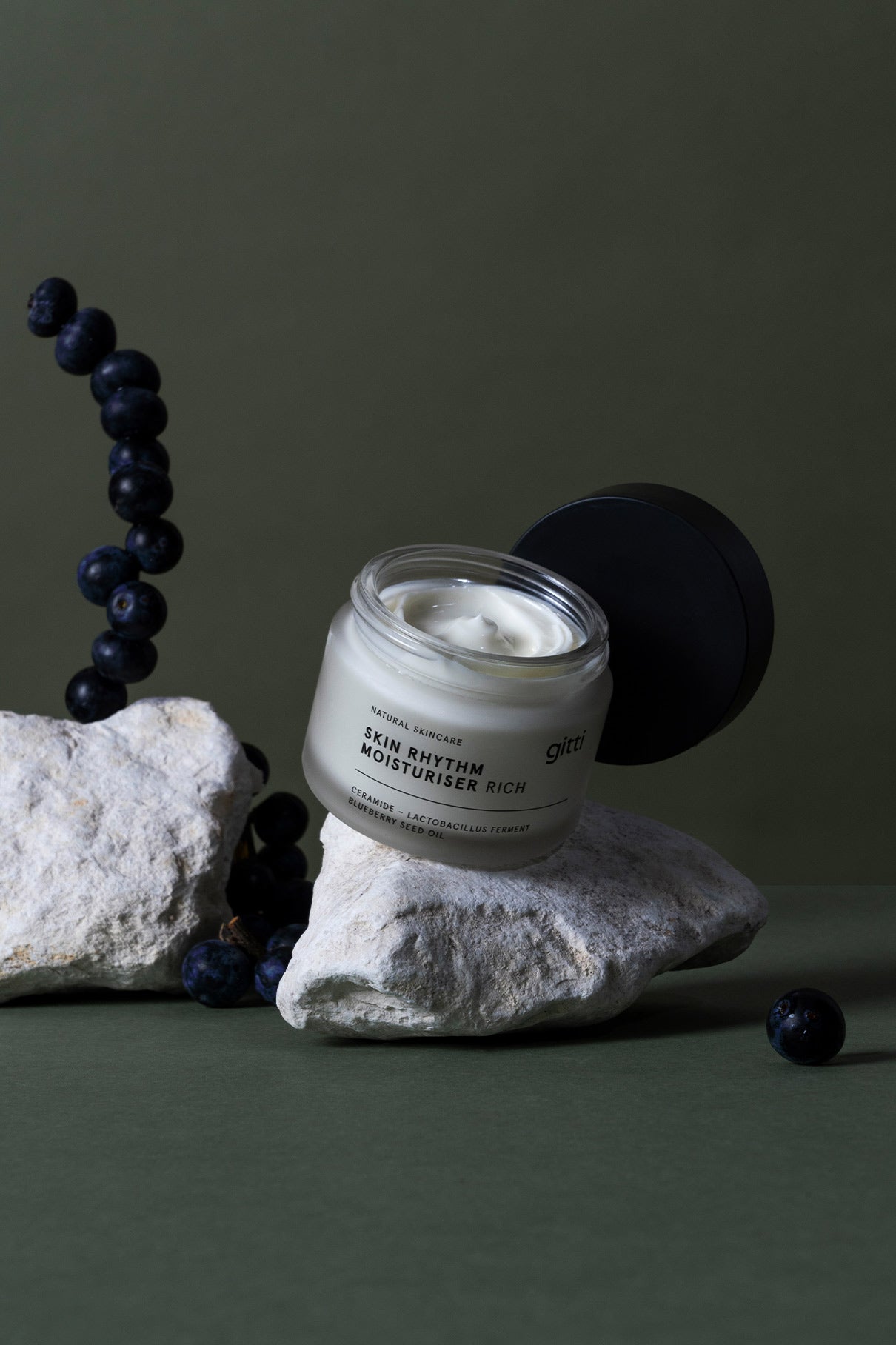 Ein geöffneter Tiegel der Essential Routine Rich Moisturiser, perfekt für die tägliche Schönheitsroutine, steht auf einem weißen Stein zwischen Blaubeeren und anderen Steinen mit grünem Hintergrund.