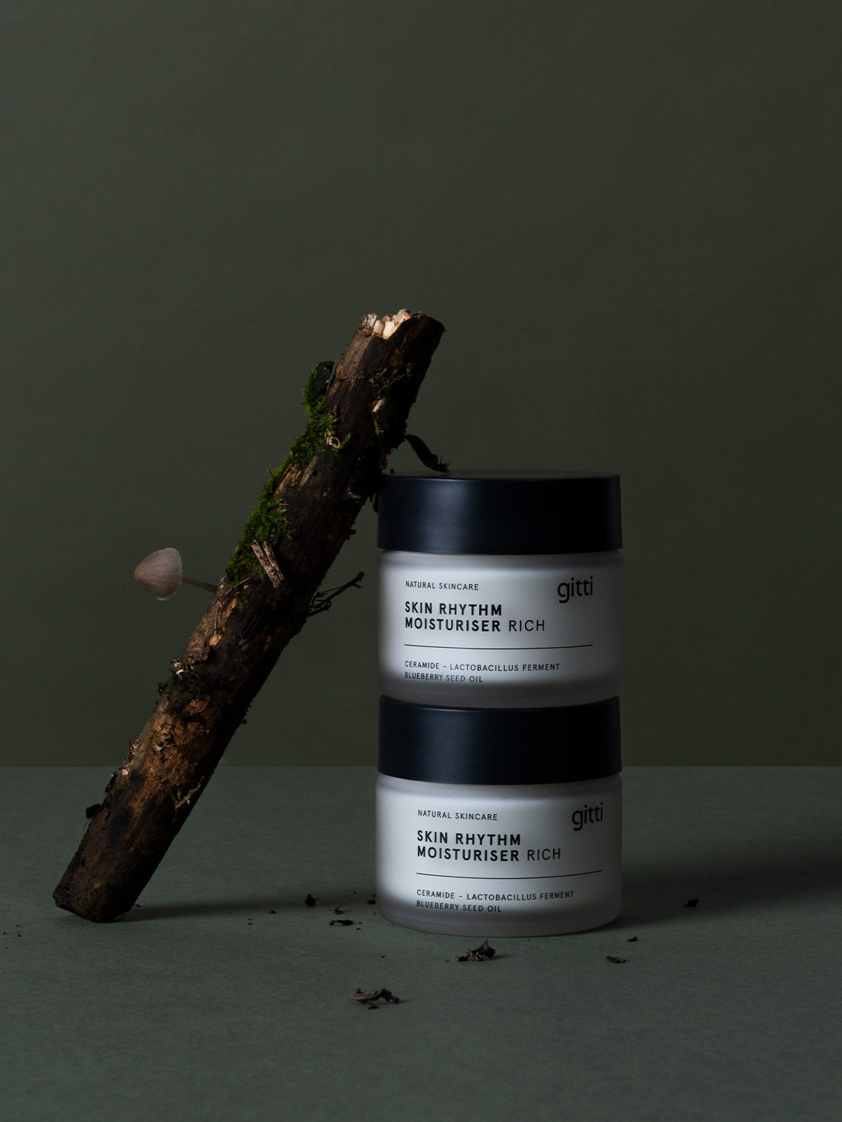 Zwei Tiegel Skin Rhythm Moisturiser Rich sind neben einem moosbewachsenen Zweig und einem Pilz gestapelt und betonen die reichhaltige Feuchtigkeit und den Barriereschutz vor einem schlichten dunklen Hintergrund.