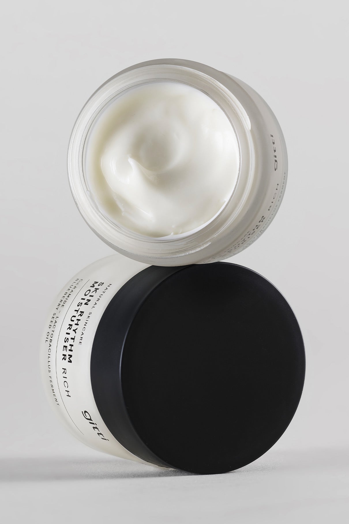 Offener Tiegel der Skin Rhythm Moisturiser Rich mit einem geschlossenen Tiegel darunter, beide in minimalistischer weißer und schwarzer Verpackung auf einfarbigem Hintergrund - für Glow, intensive Feuchtigkeit und Barriereschutz konzipiert.