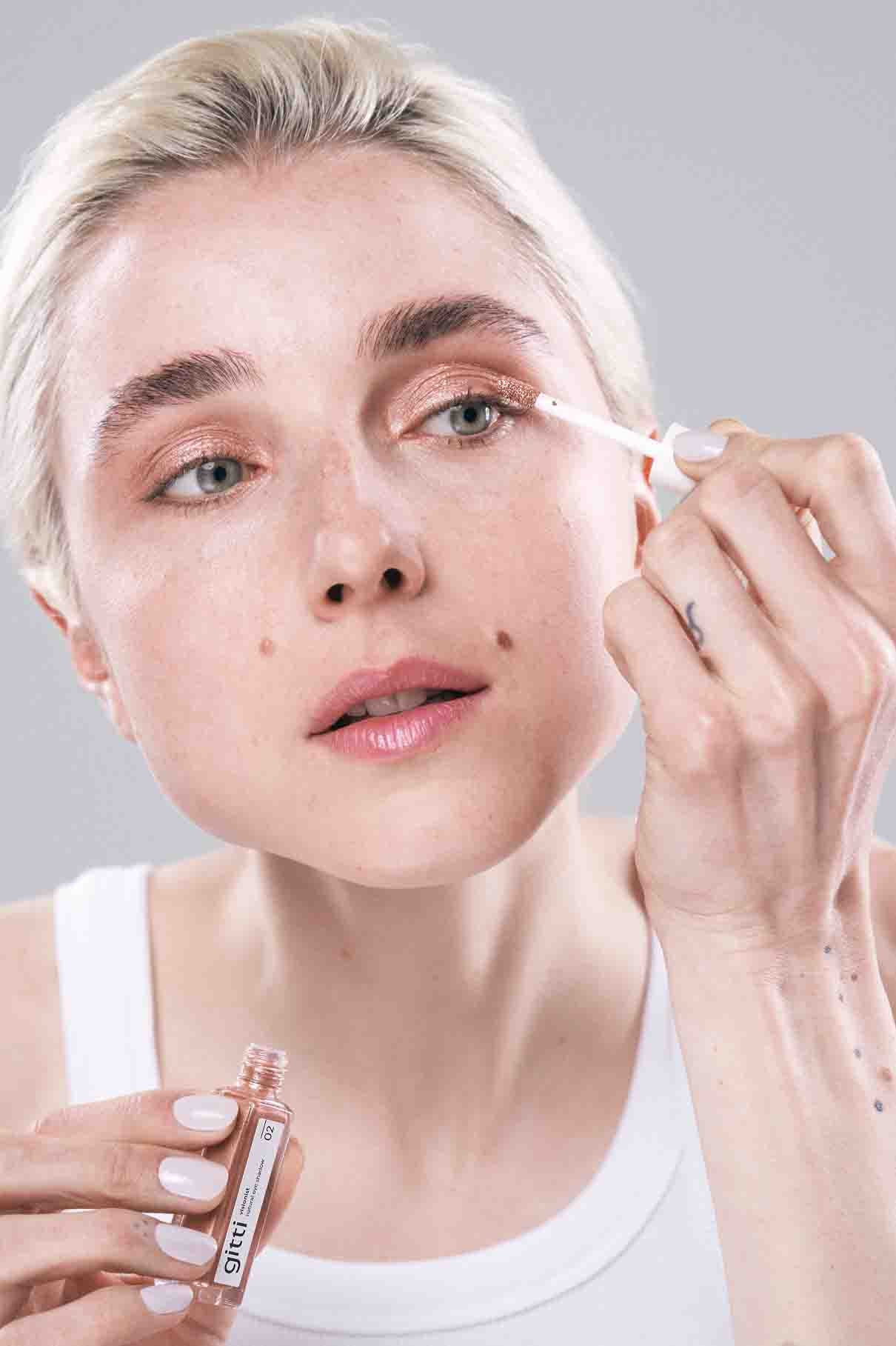 Eine Person trägt einen schimmernden Lidschatten aus dem Visionist Celebration Duo mit einem Applikator auf ihr Augenlid auf, während sie ein kleines Make-up-Fläschchen hält und ein weißes Oberteil trägt.