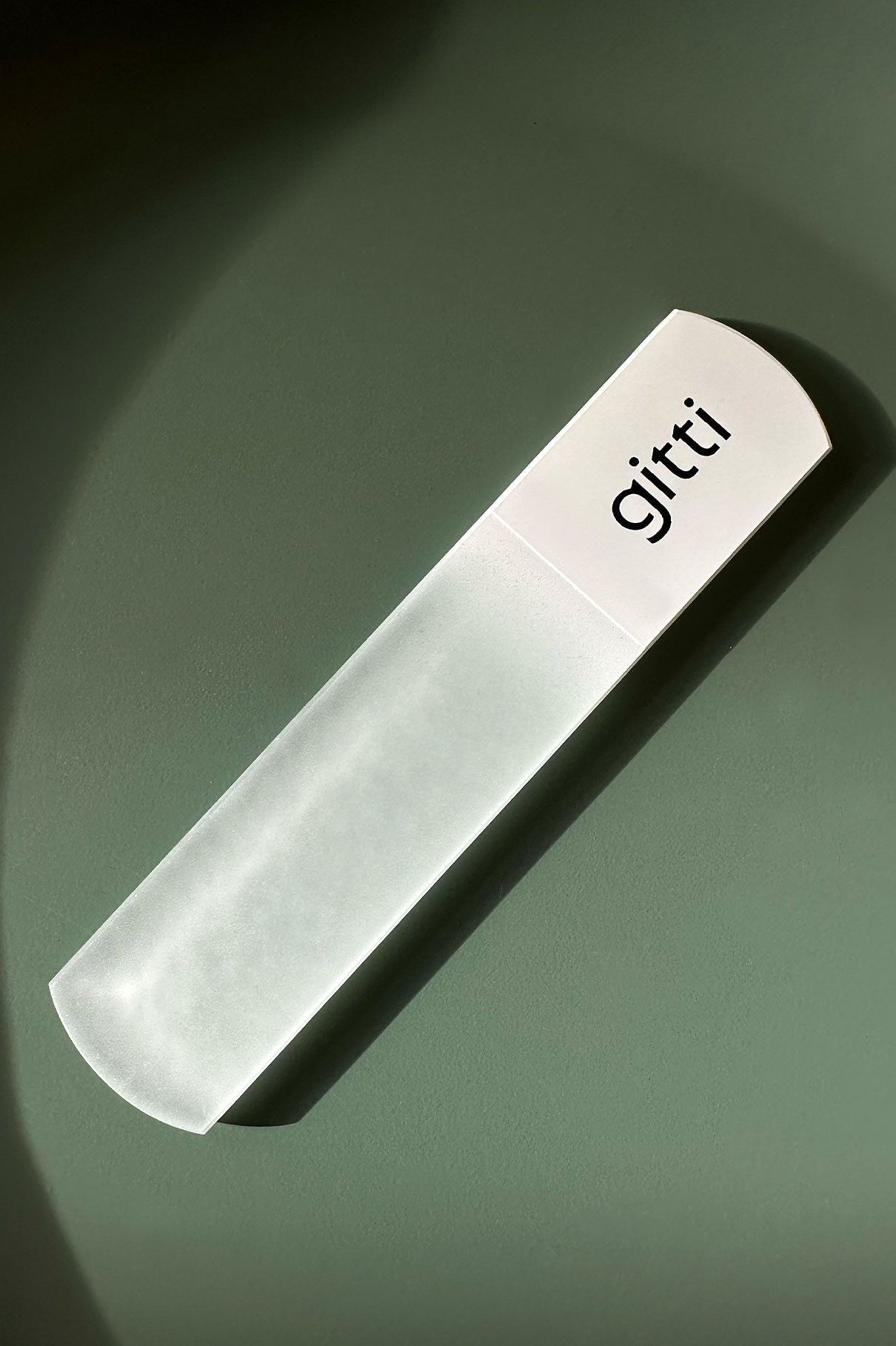 Eine Fußfeile mit einer mattierten Glasoberfläche und einem weißen Griff mit der Aufschrift gitti ruht auf einem flachen, gedämpften grünen Hintergrund - ideal für die sanfte Fußpflege.