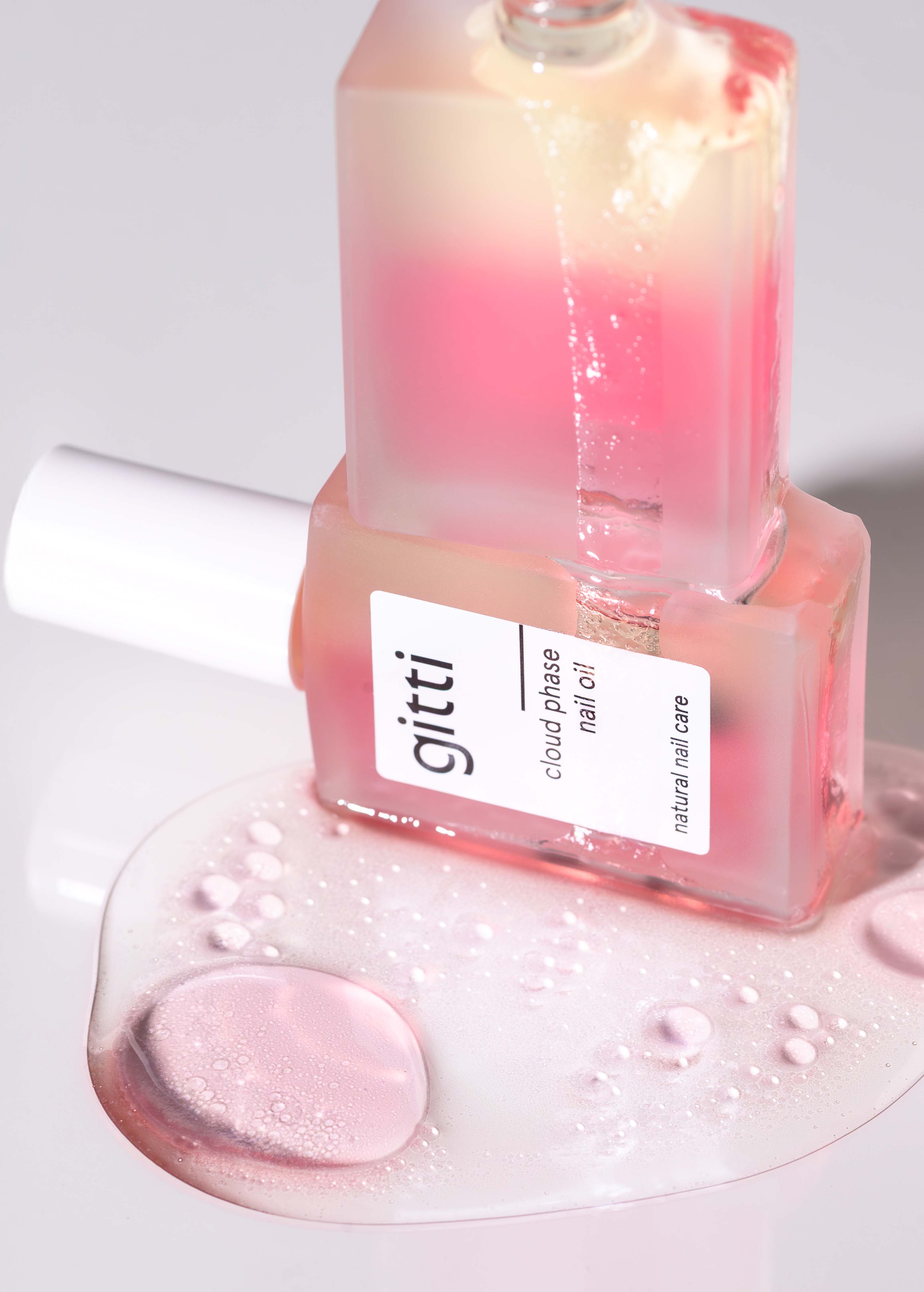 Zwei Glasflaschen mit gitti Cloud Phase Nagelöl, eine aufrecht und eine liegend mit verschüttetem rosa Öl und Tropfen auf einer reflektierenden Oberfläche - eine unverzichtbare Ergänzung zu Ihrem Perfect Nails Starter Set für wunderschöne Nägel.