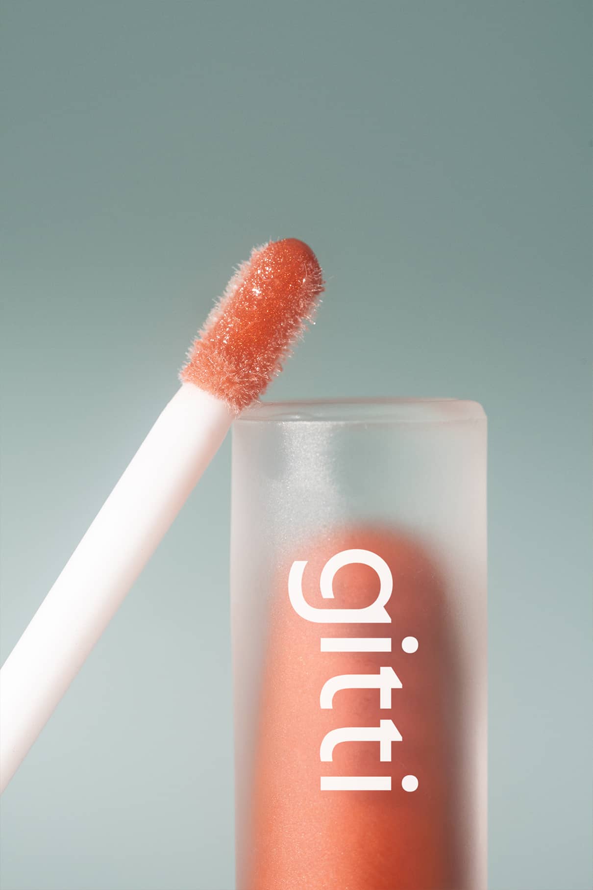 Eine Nahaufnahme des Lip Glaze no. 04 Applikator mit Nude-Ton und Hochglanz-Finish, neben einer mattierten Tube mit der Aufschrift gitti auf hellblauem Hintergrund.