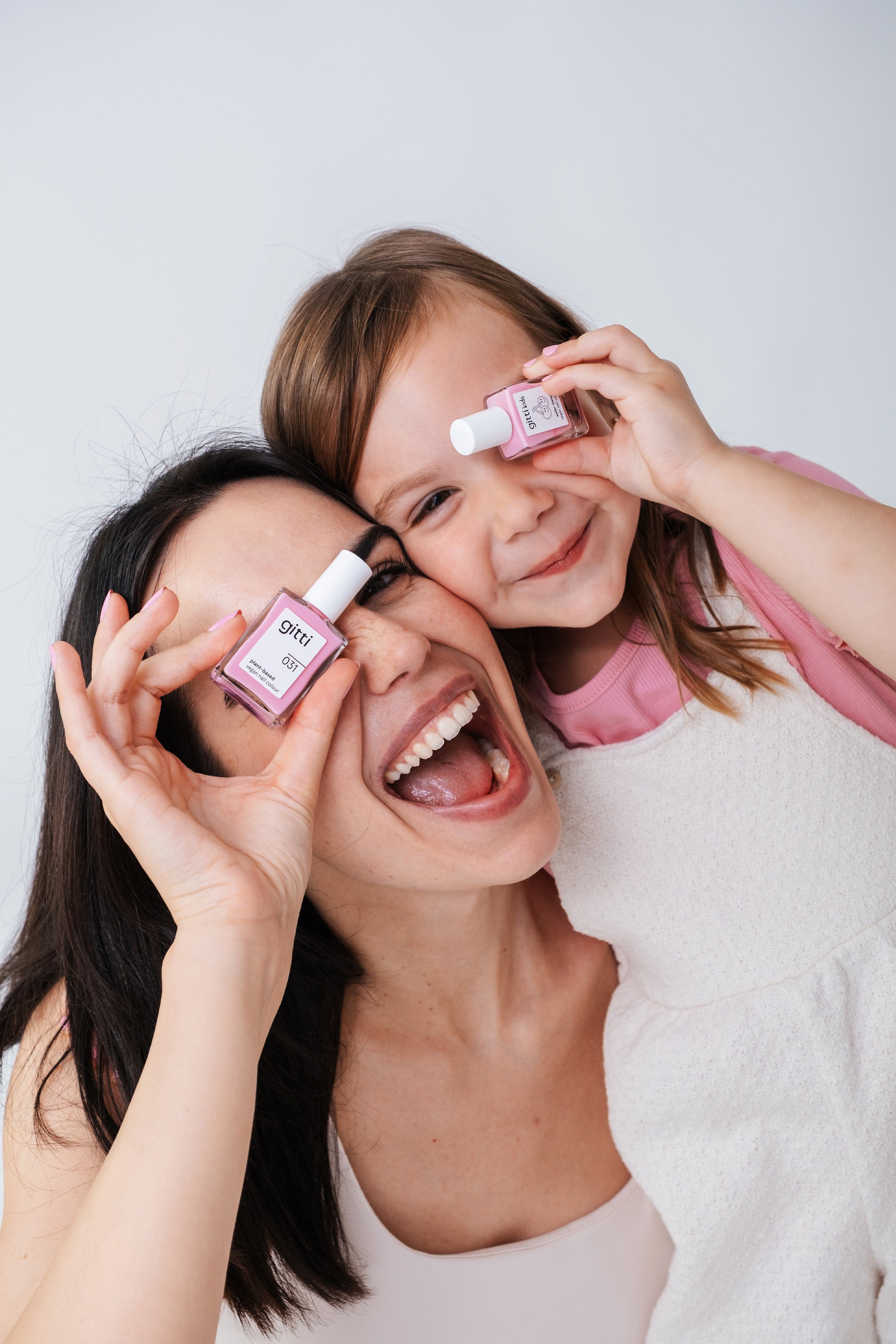 Eine Frau und ein junges Mädchen lächeln und halten spielerisch die Nagellackflaschen des Mini & Me Sets an ein Auge und posieren vor einem einfachen Hintergrund.