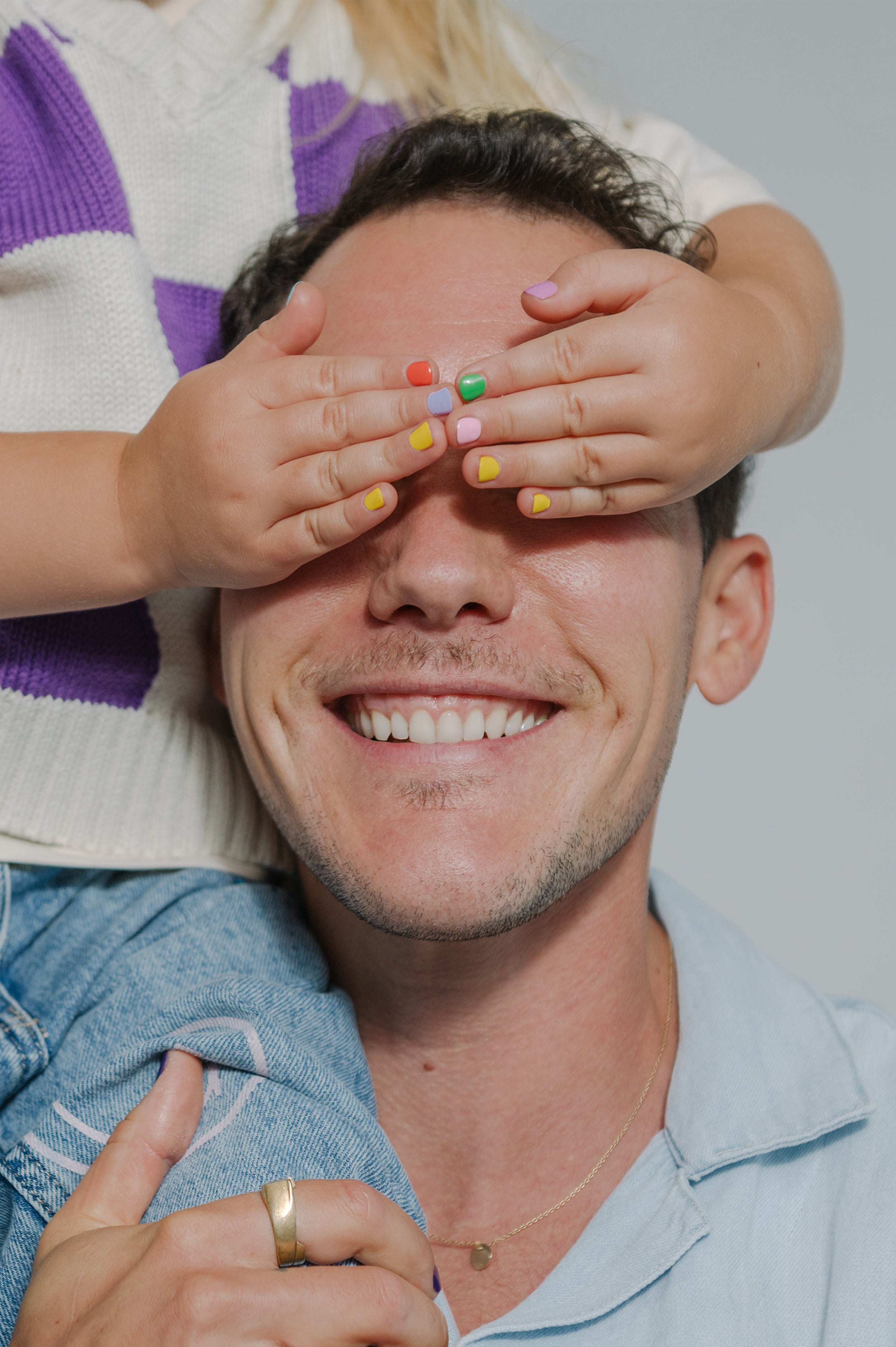 Ein lächelnder Mann wird spielerisch von Kinderhänden mit buntem Kids Duo Nagellack auf den Augen bedeckt. Das Kind, das auf seinen Schultern sitzt, zeigt seine kinderfreundlichen Kids Duo-Nägel.