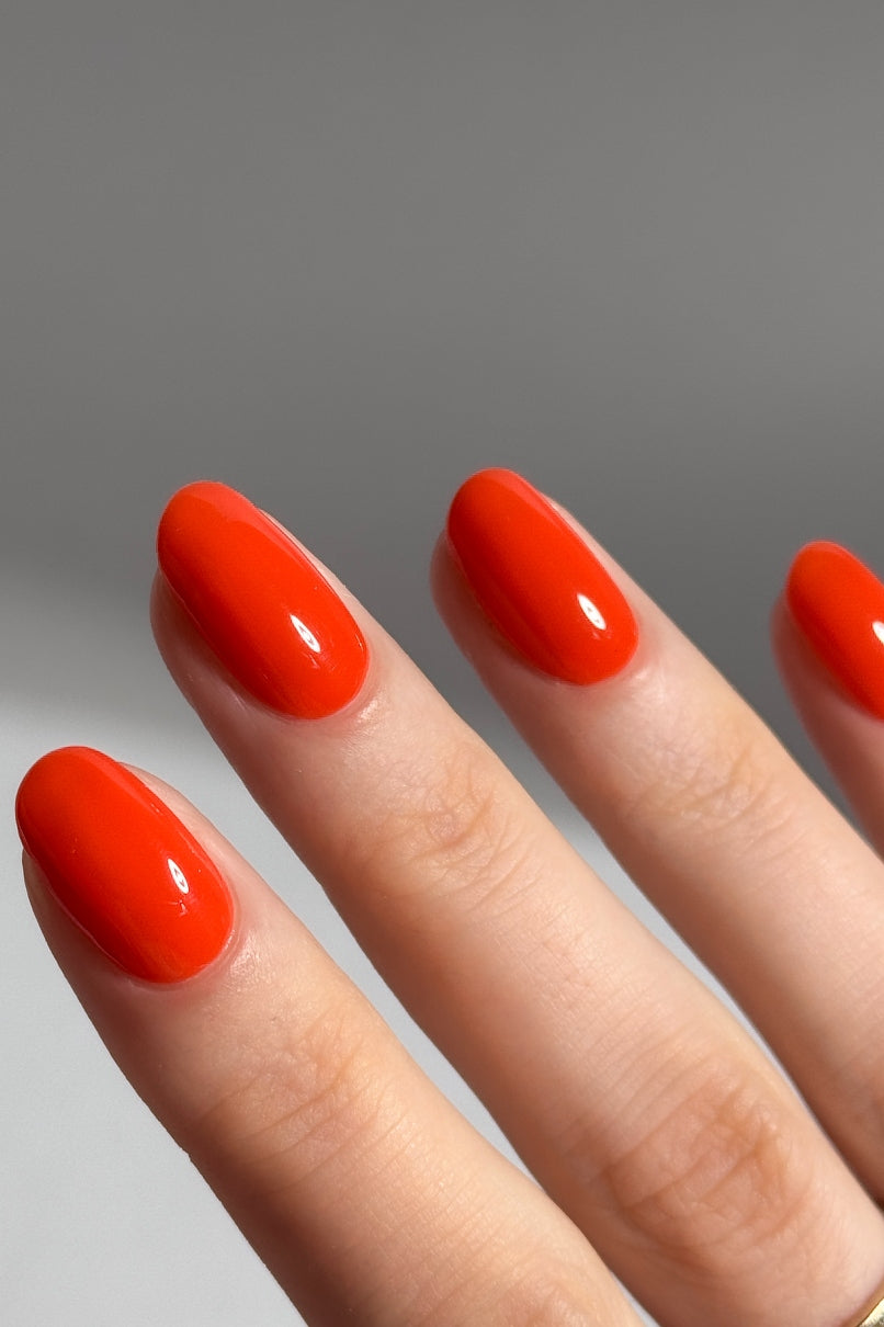 Eine Nahaufnahme einer Hand mit sauber manikürten Nägeln, die mit glänzendem, leuchtend rot-orangem Lack lackiert und mit dem plantPRO Top Coat veredelt wurden, zeigt einen gelartigen Glanz vor einem schlichten Hintergrund.