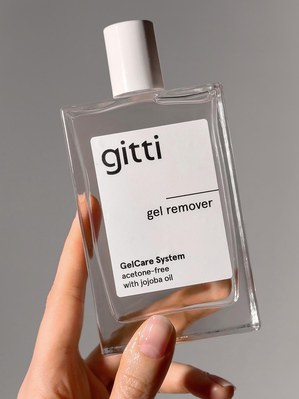 Eine Hand, die eine durchsichtige, rechteckige Flasche mit dem acetonfreien GelCare Remover mit Jojobaöl hält.