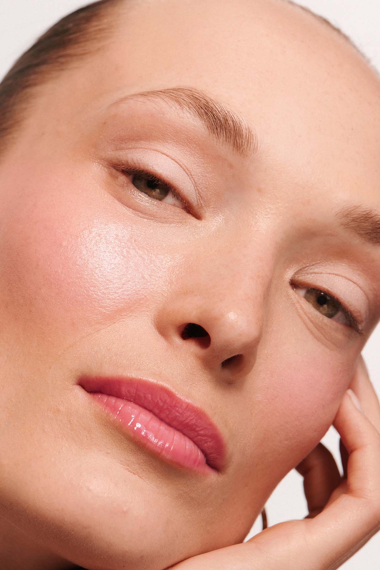 Nahaufnahme des Gesichts einer Person mit reiner Haut, natürlichem Make-up mit dem „Velvet Cheeks Nr. 03 silikonfrei Mousse-Blush“ in Pastellrosa, rosa Lippen und einem entspannten Gesichtsausdruck, wobei die Person ihre Wange auf die Hand stützt.