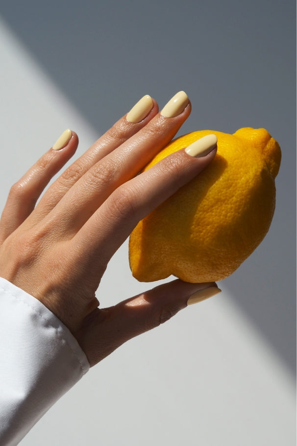 Eine Hand mit hellgelb manikürten Nägeln, lackiert mit Quick Manicure Duo pflanzenbasierte Farbe, hält eine gelbe Zitrone vor einem weißen Hintergrund mit Schatten.