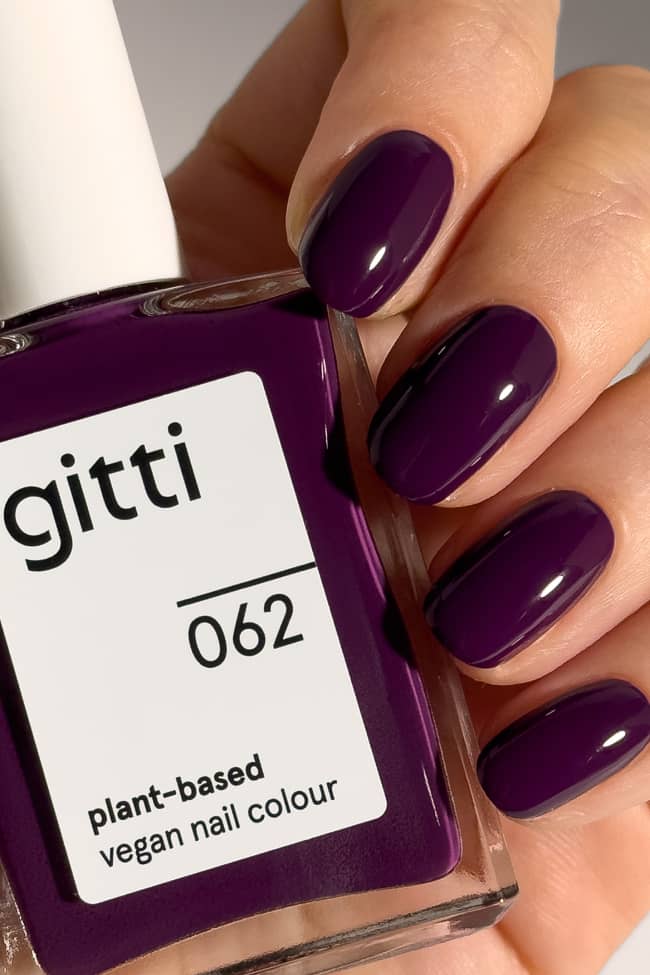 Eine Hand mit kurzen, abgerundeten Nägeln, die in einem glänzenden, tiefen Violett mit rötlichem Unterton lackiert sind, hält eine Flasche des pflanzlichen, veganen Nagellacks „Gitti Nr. 062“ in genau demselben pflaumenvioletten Farbton.