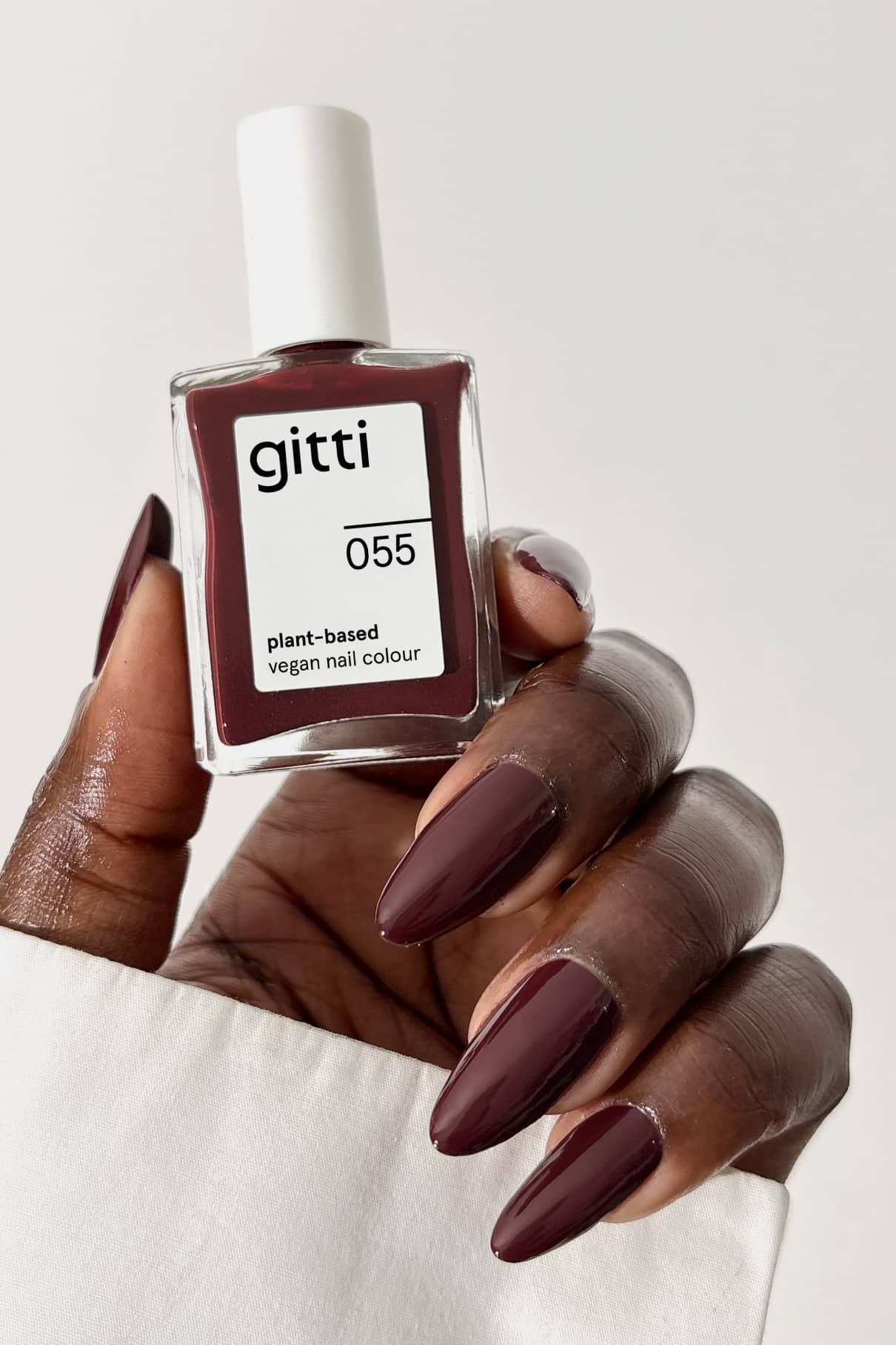 Eine Hand mit dunkelkastanienbraunen, mahagonilackierten Nägeln hält eine Flasche gitti no. 055 - eine pflanzliche, vegane Nagelfarbe in einem von Espresso und Martini inspirierten Farbton - vor einem schlichten Hintergrund.