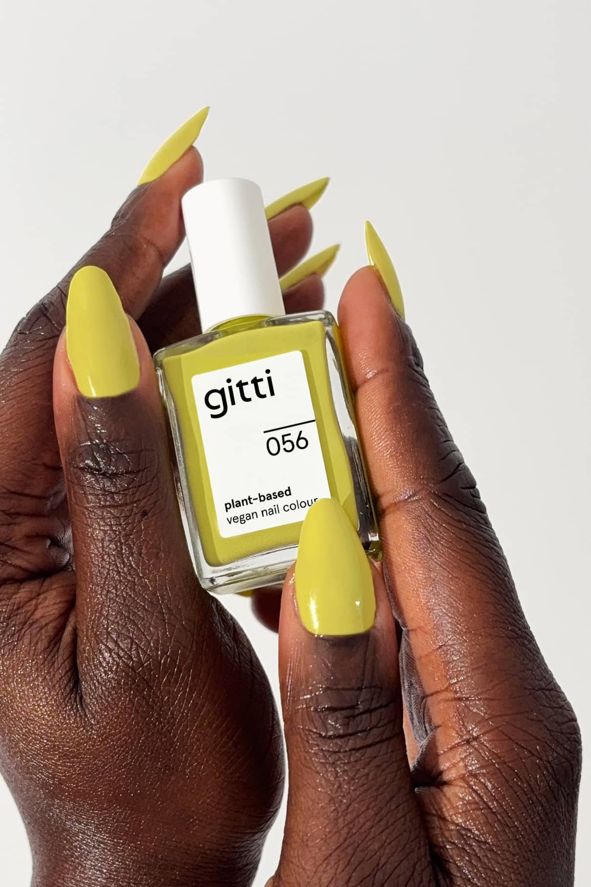 Eine Person mit gelb-grün lackierten Nägeln, wie gitti no. 056, hält eine Flasche vegane Nagelfarbe auf Pflanzenbasis vor einem weißen Hintergrund.