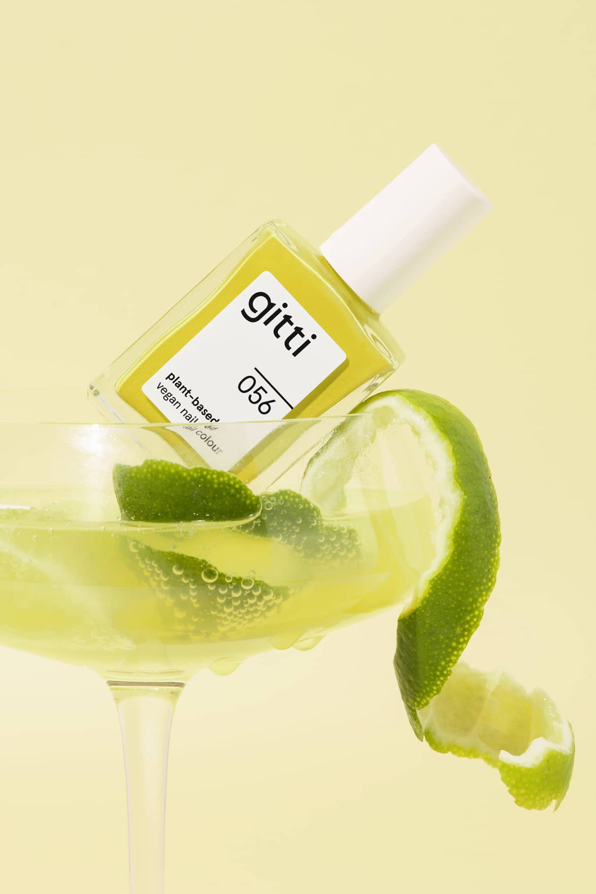 Eine Flasche gitti no. 056 Nagellack befindet sich in einem Coupe-Glas, das mit zitrusgelber Flüssigkeit und Limettenschalen gefüllt ist, vor einem gelben Hintergrund.