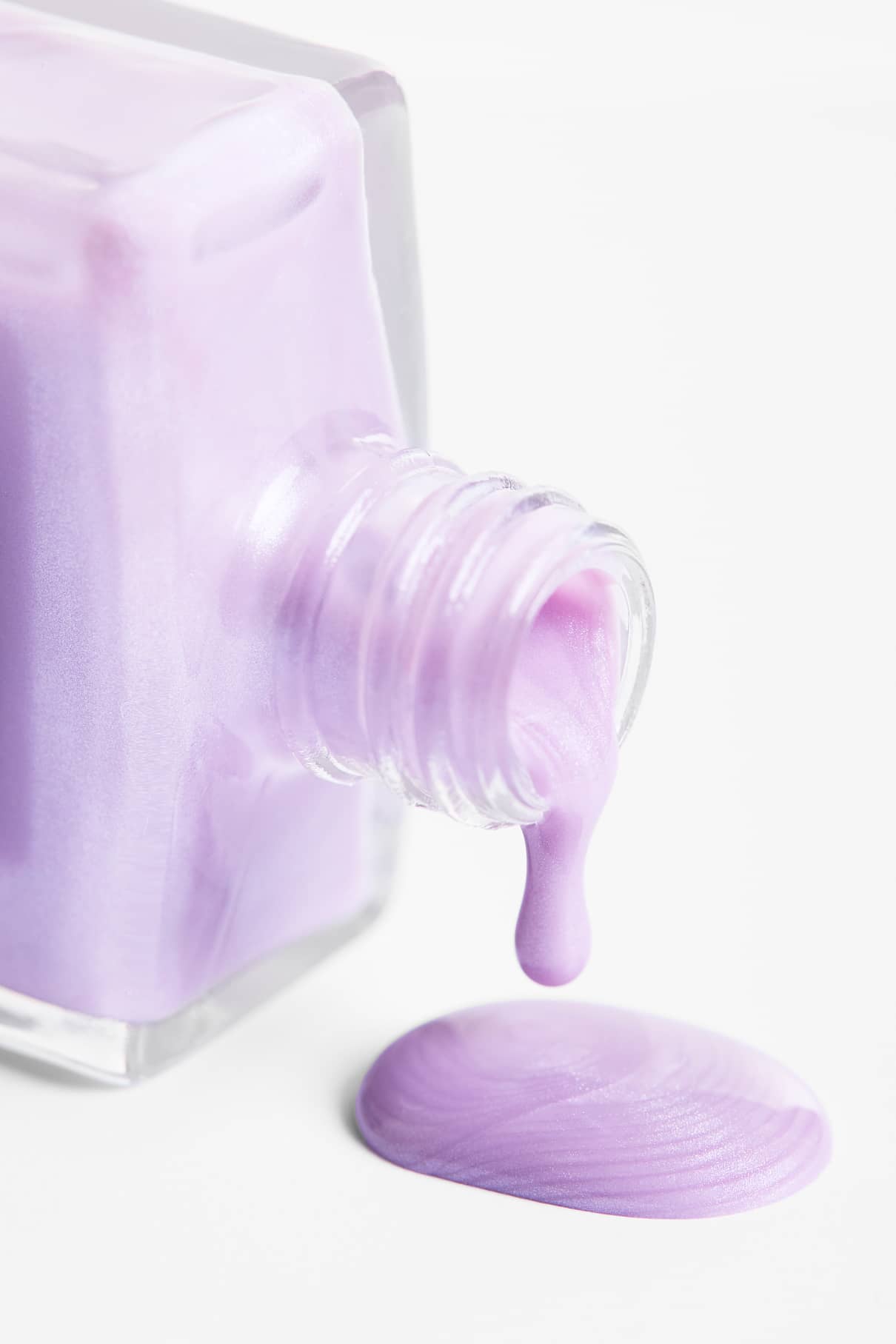 Ein Glasfläschchen mit Skinified Nail Tint 04 (hellviolett) ist umgekippt, wodurch der Lack verschüttet wurde und sich eine kleine Pfütze auf einer weißen Fläche gebildet hat.