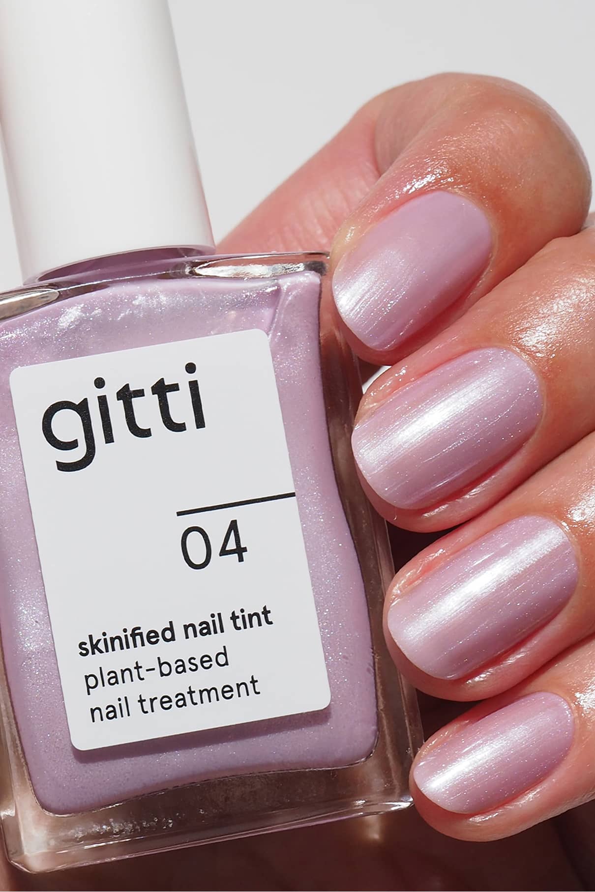 Eine Hand mit kurzen, sauber manikürten Nägeln, die in einem schimmernden Hellrosa lackiert sind, hält eine Flasche Skinified Nail Tint 04.