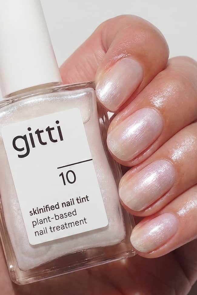 Eine Hand mit kurzen, natürlichen Nägeln, die in einem schimmernden, perlmuttartigen Farbton lackiert sind, hält eine 15-ml-Flasche mit der Aufschrift gitti 10-part des pflanzlichen Treatment Duo Nail Treatment, das perfekt für jedes Selbstpflegeset ist.