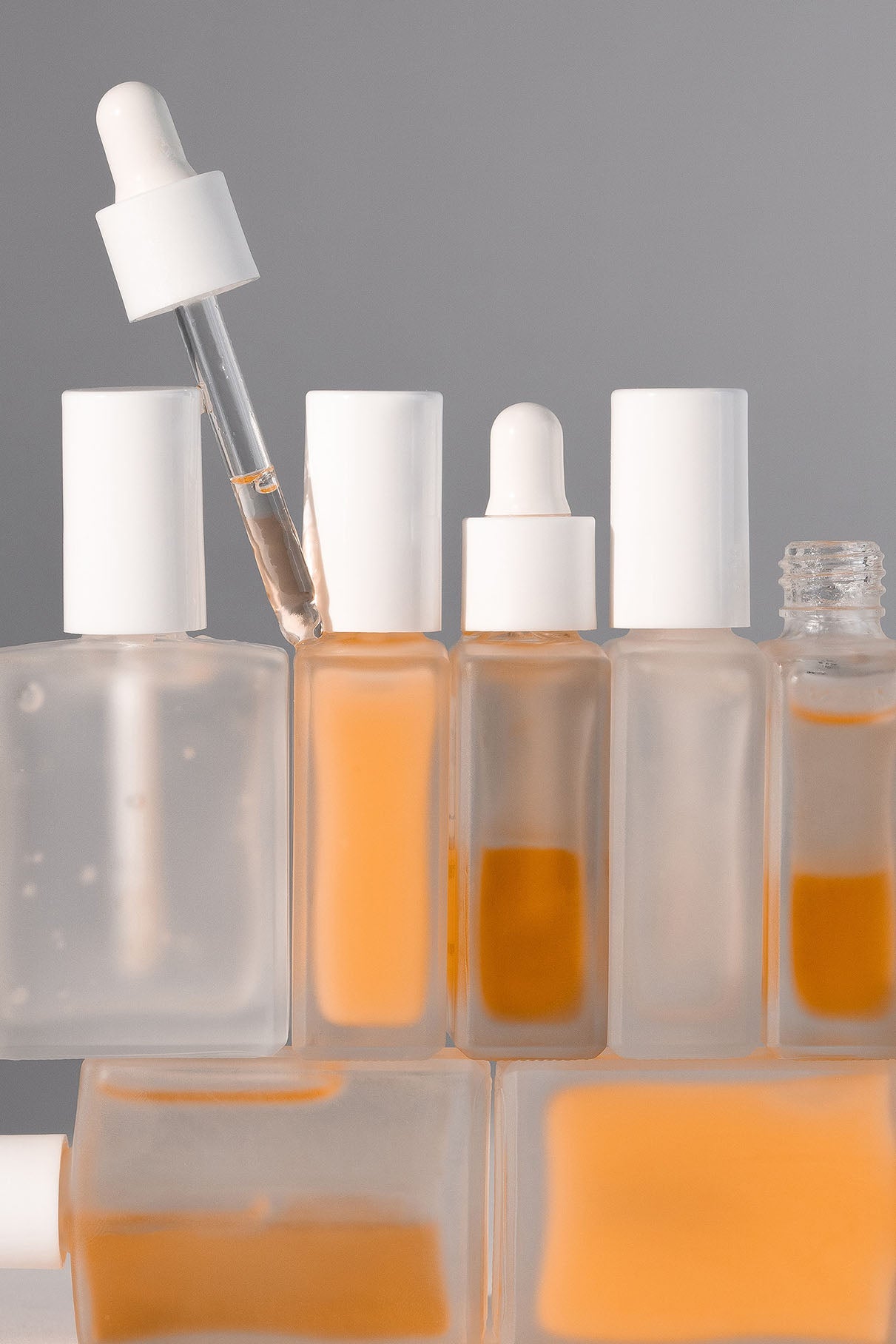 Eine Gruppe von Milchglas-Tropfflaschen, gefüllt mit orangefarbener Flüssigkeit, einige offen und einige geschlossen, in Reihen vor einem schlichten grauen Hintergrund angeordnet - perfekt für Ihre Nagelpflege: Nail Recovery Treatment mit natürlichen Inhaltsstoffen.