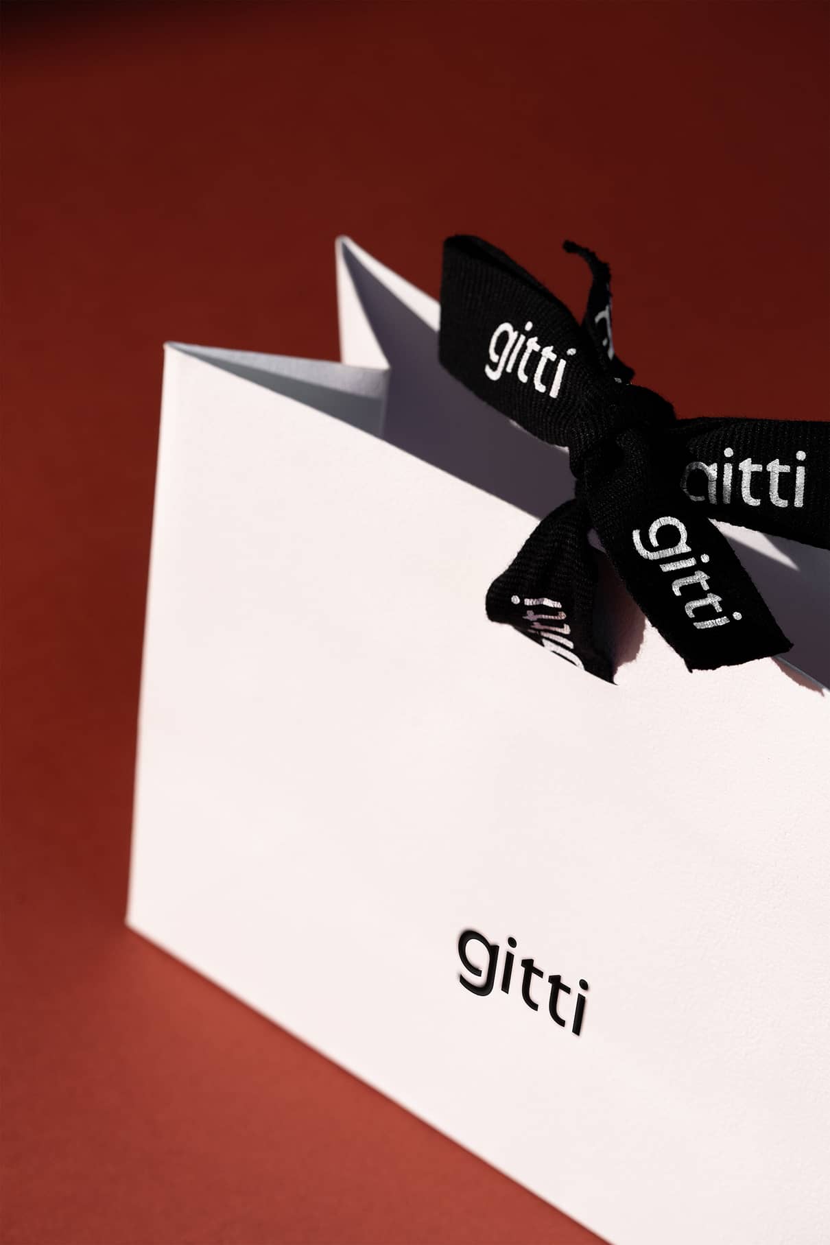 Eine weiße Geschenktüte mit dem Aufdruck "gitti", mit einer schwarzen Schleife und einem Etikett, auf dem ebenfalls "gitti" steht, vor einem rotbraunen Hintergrund.