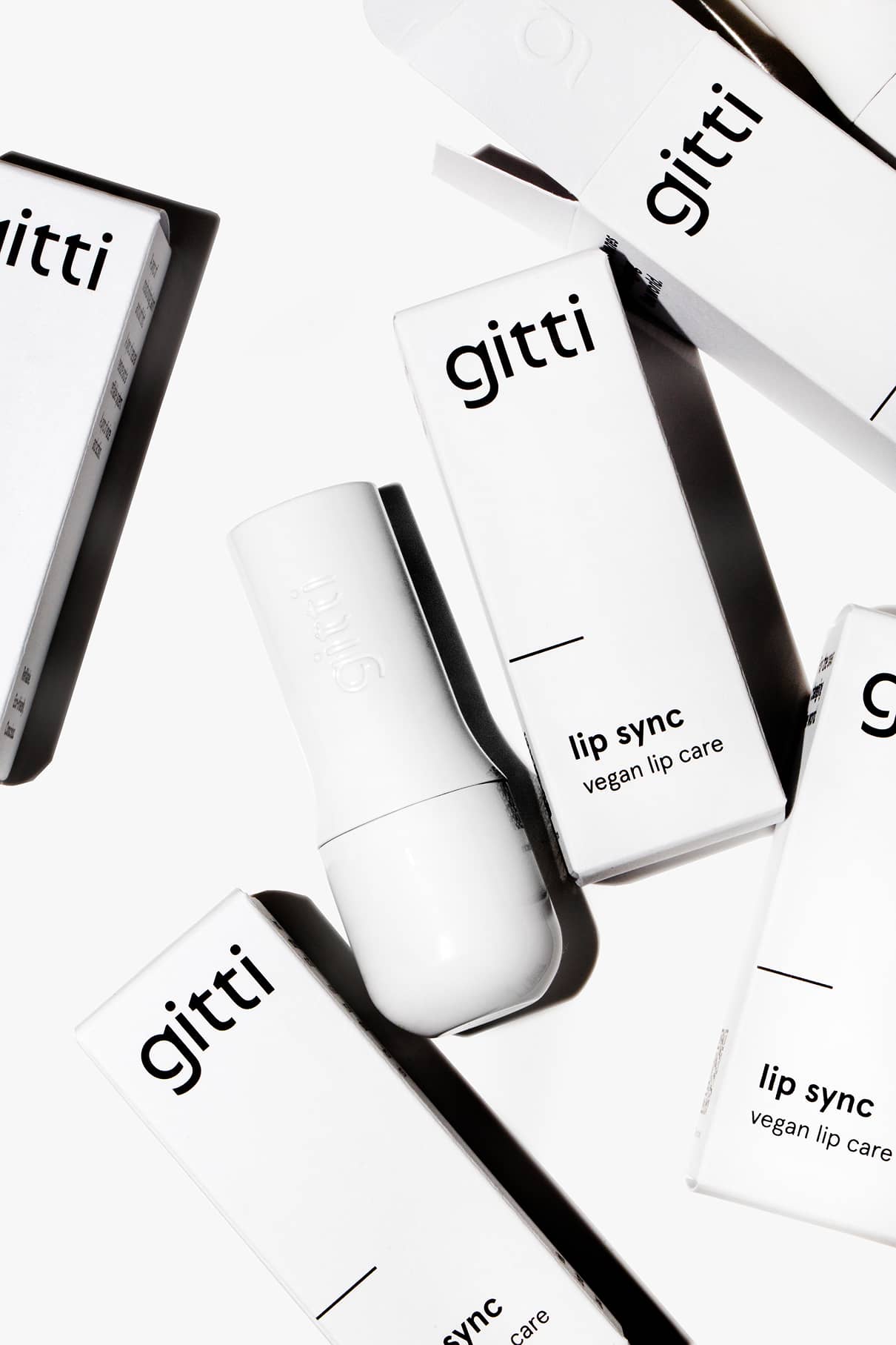 Mehrere weiße Schachteln mit der Aufschrift gitti Lip Sync vegane Lippenpflege sind um ein weißes Lip Sync Case mit einem nachfüllbaren Deckel auf weißem Hintergrund angeordnet.