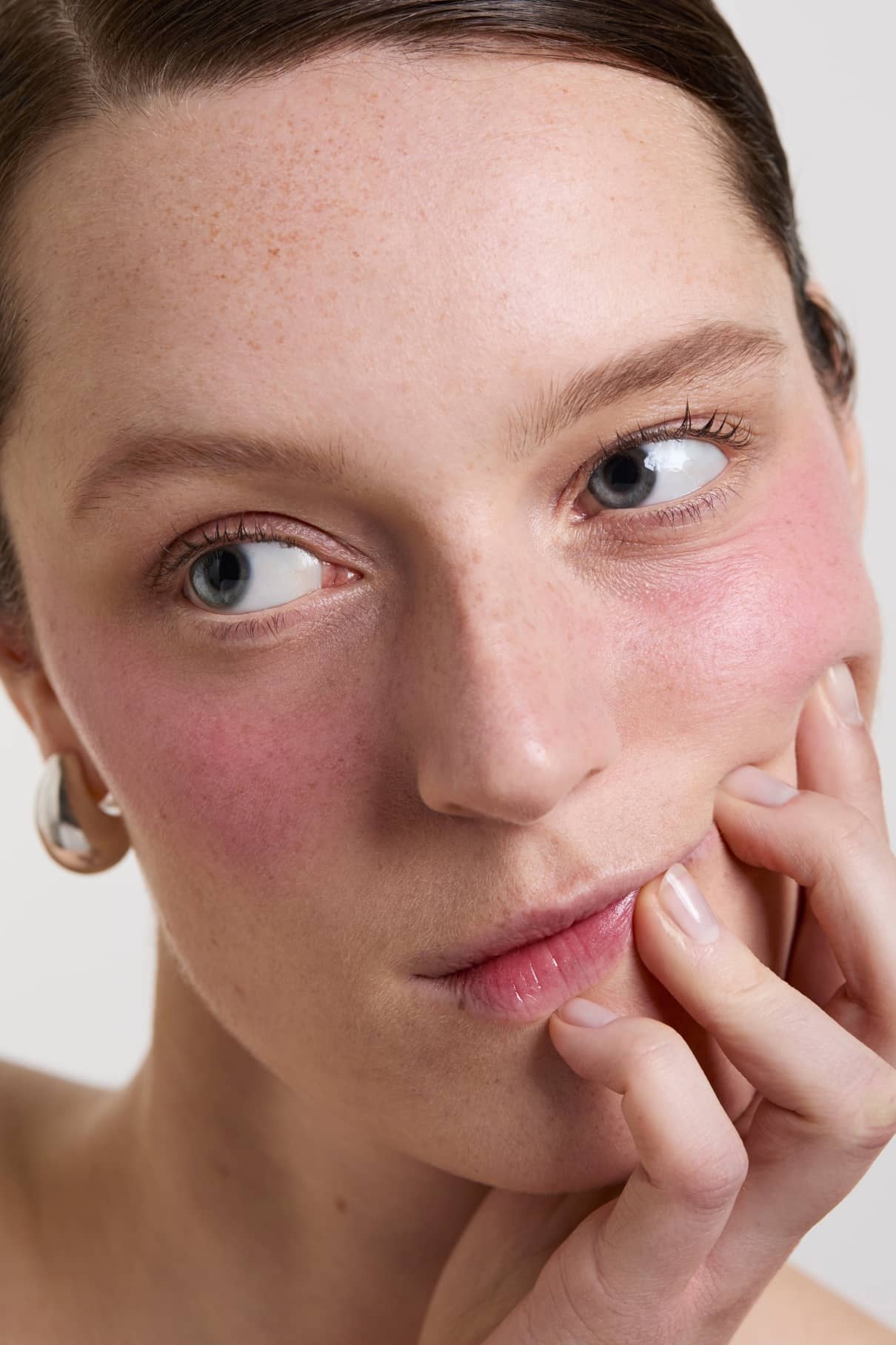 Nahaufnahme einer Person mit heller Haut und blauen Augen, die zur Seite schaut, während sie ihr Gesicht berührt. Ihre geröteten Wangen werden durch Velvet Cheeks no. 05 für einen frischen, silikonfreien Look.