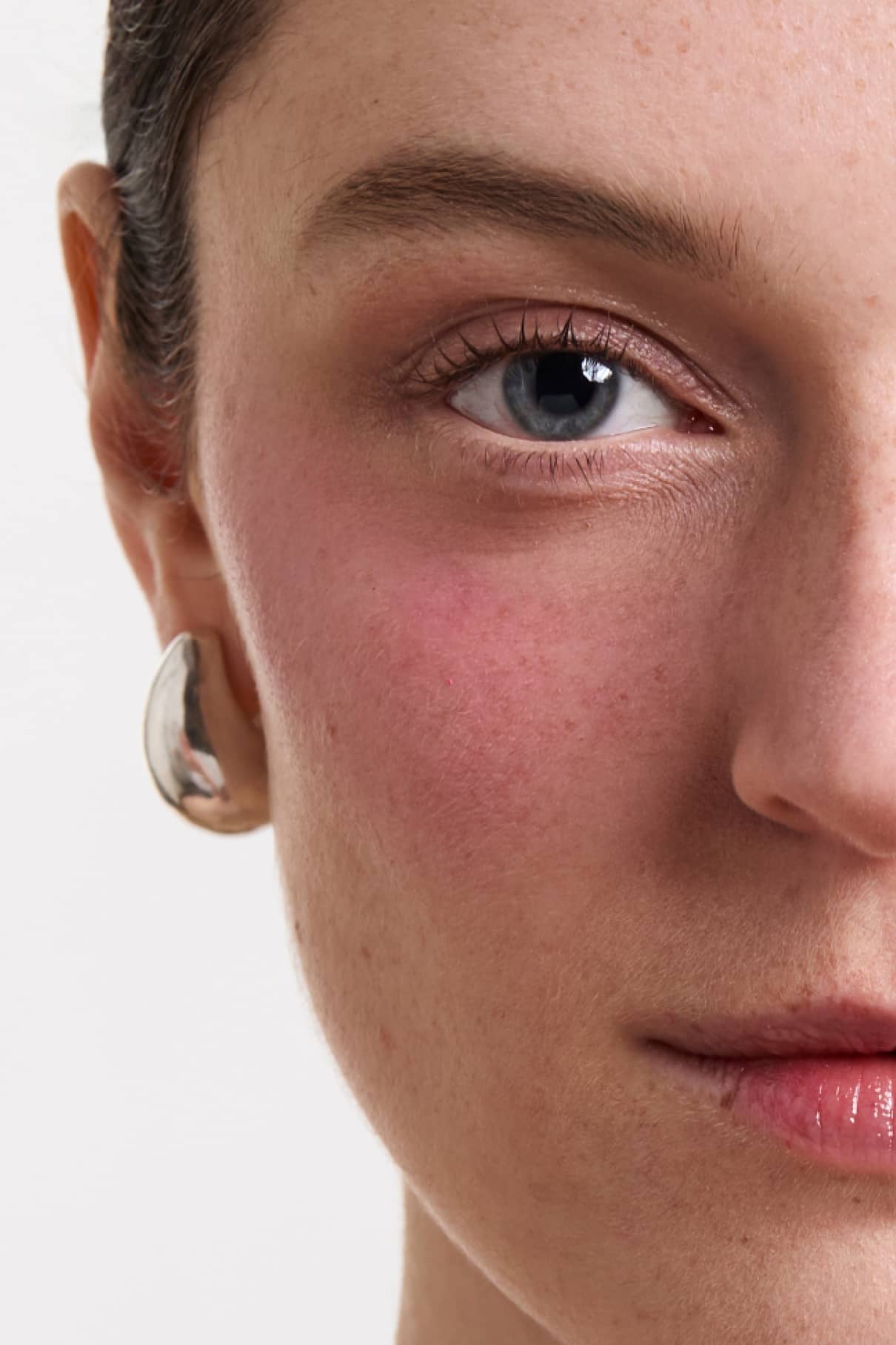 Nahaufnahme einer Frau mit heller Haut, minimalem Make-up und einem metallischen Ohrring. Ein Auge ist mit der Wimperntusche Lash & Cheek Duo umrandet und die Wangen sind samtweich geschminkt; ein Teil der Lippen ist ebenfalls zu sehen.
