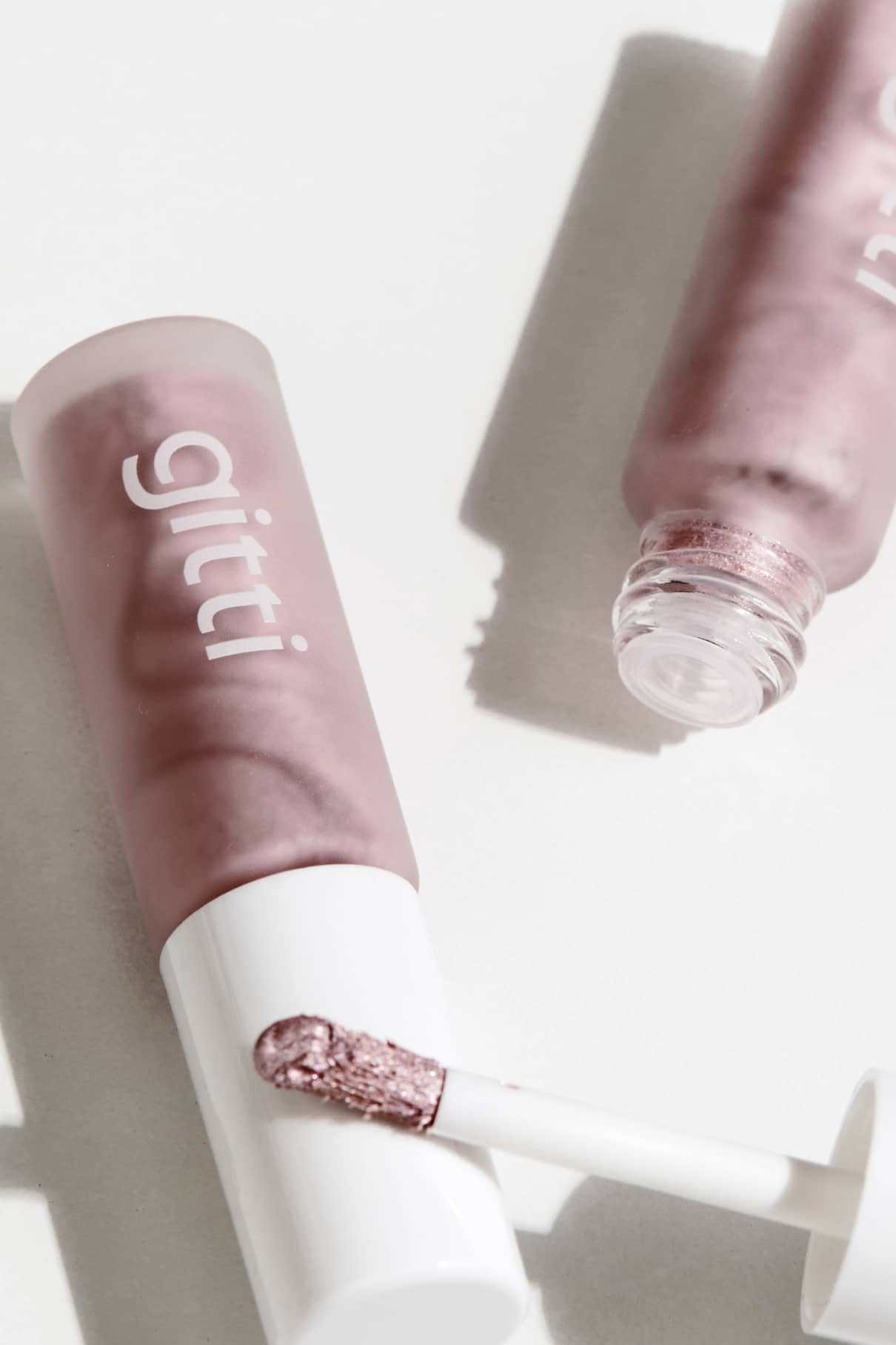 Zwei mauvefarbene Halo Eyes no. 04 Lipgloss-Tuben, von denen eine geöffnet ist, um ihren schimmernden, lichtreflektierenden Rosé-Ton zu enthüllen, sind auf einer weißen Fläche mit weichen Schatten platziert.