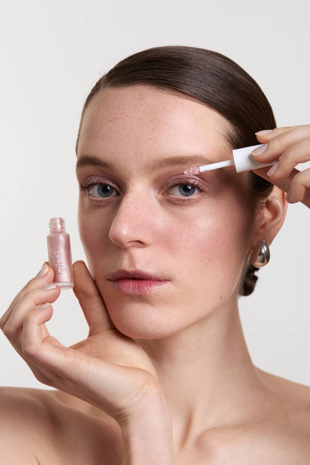 Eine Frau trägt Halo Eyes no. 04, ein schimmernder, lichtreflektierender roséfarbener Lidschatten, wird mit einem Pinselapplikator auf ihr Augenlid aufgetragen, wobei sie das Produkt nahe an ihr Gesicht hält.