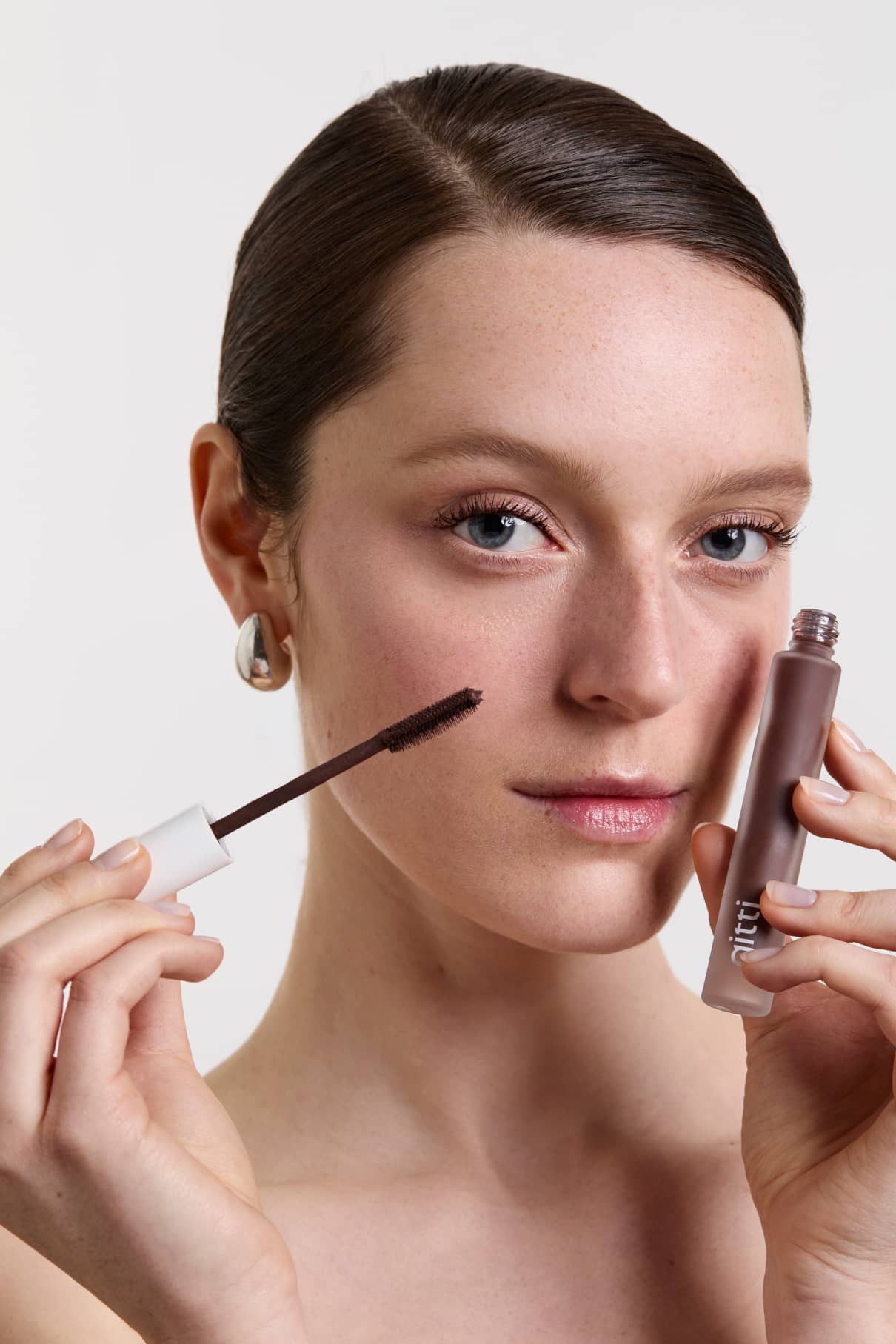 Eine Frau mit heller Haut hält einen Infinite Lash Nr. 02-Zauberstab in die Nähe ihres Gesichts und zeigt die Wimperntusche-Tube, wobei sie direkt in die Kamera vor einem unifarbenen Hintergrund blickt.