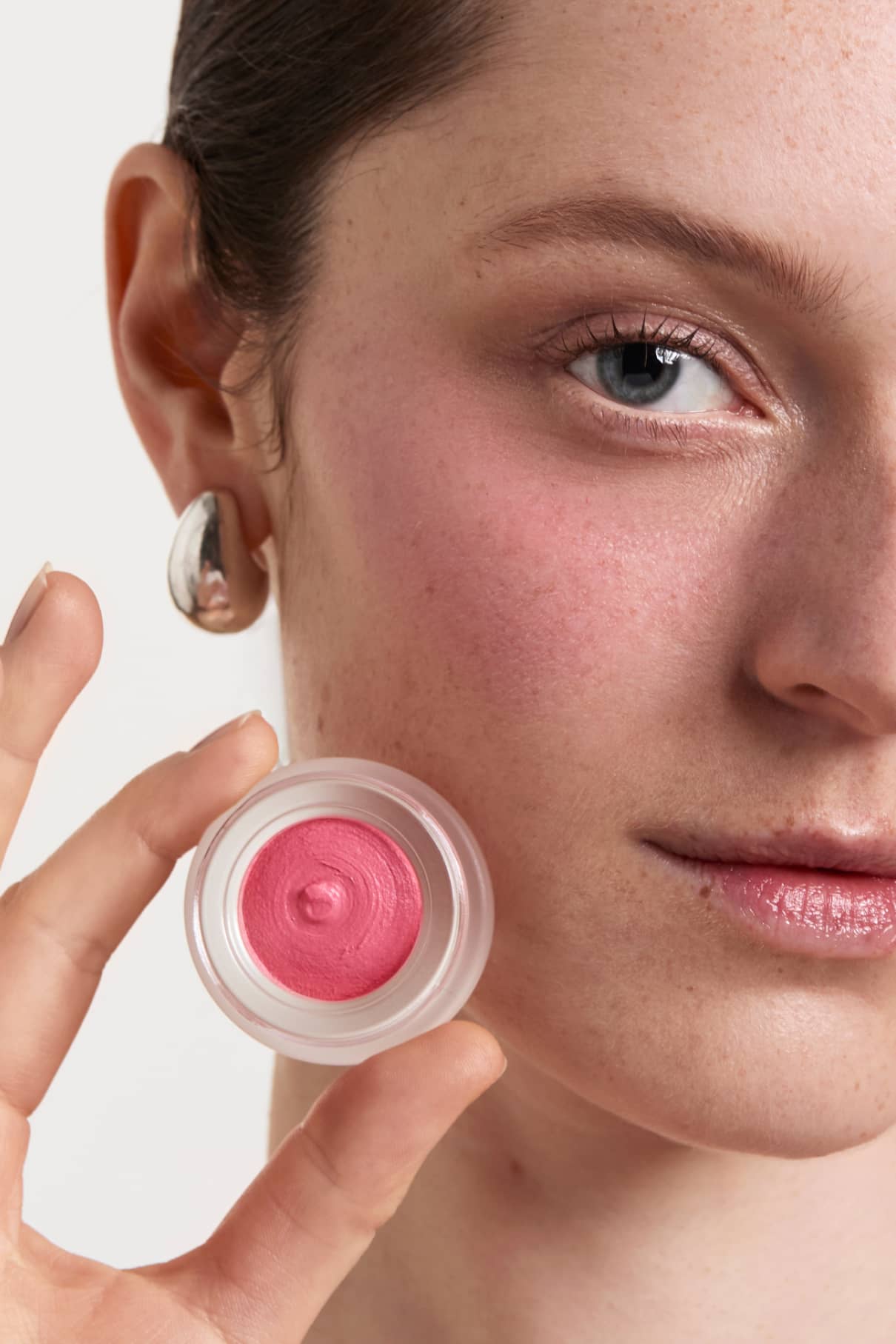 Eine Person hält eine offene Dose Velvet Cheeks Nr. 05, ein rosafarbenes silikonfreies Mousse-Blush, in die Nähe ihres Gesichts, wobei das Rouge auf ihre Wange aufgetragen wird.