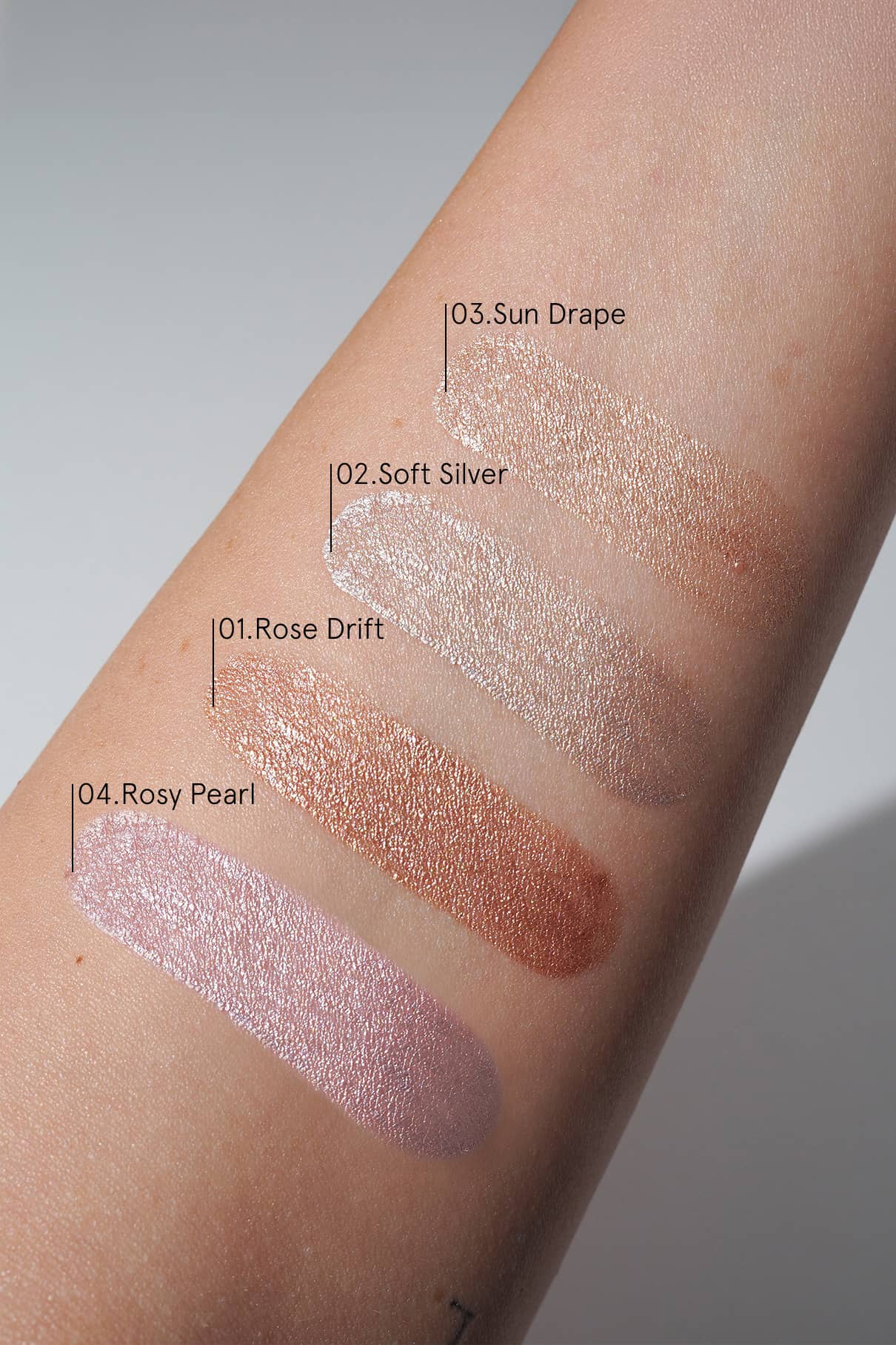 Vier schimmernde Halo Eyes no. 04 Lidschatten - Rose Drift, Soft Silver, Sun Drape und Rosy Pearl - werden auf einem Unterarm vor einem neutralen Hintergrund gezeigt, um ihr lichtreflektierendes Finish und ihre sanften Rosatöne zu betonen.