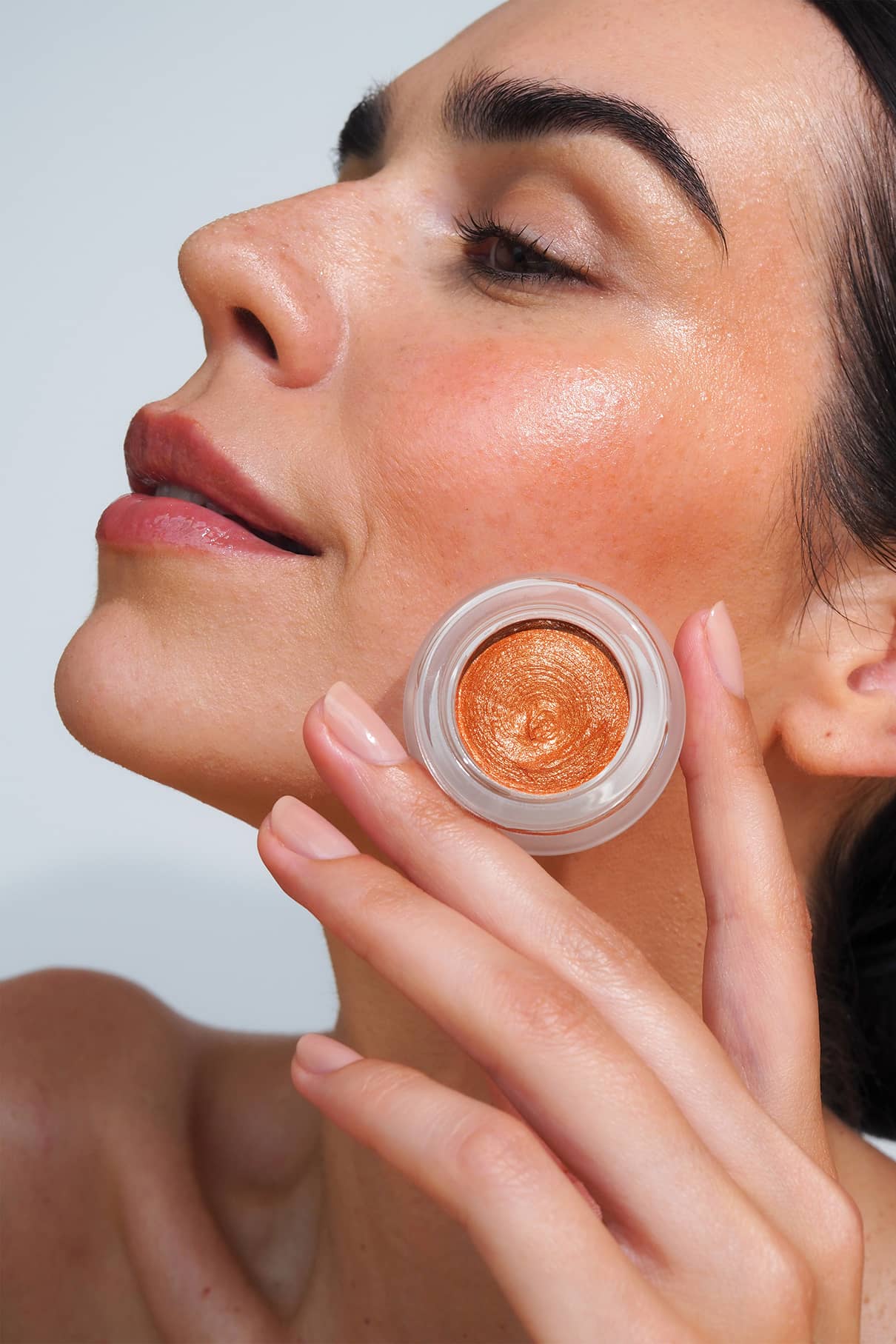 Eine Person mit strahlender Haut hält ein offenes Töpfchen mit schimmernder, bronzefarbener Creme-Make-up-Creme nahe an ihre Wange und präsentiert damit „Velvet Cheeks Nr. 04“ für ein natürliches Finish.