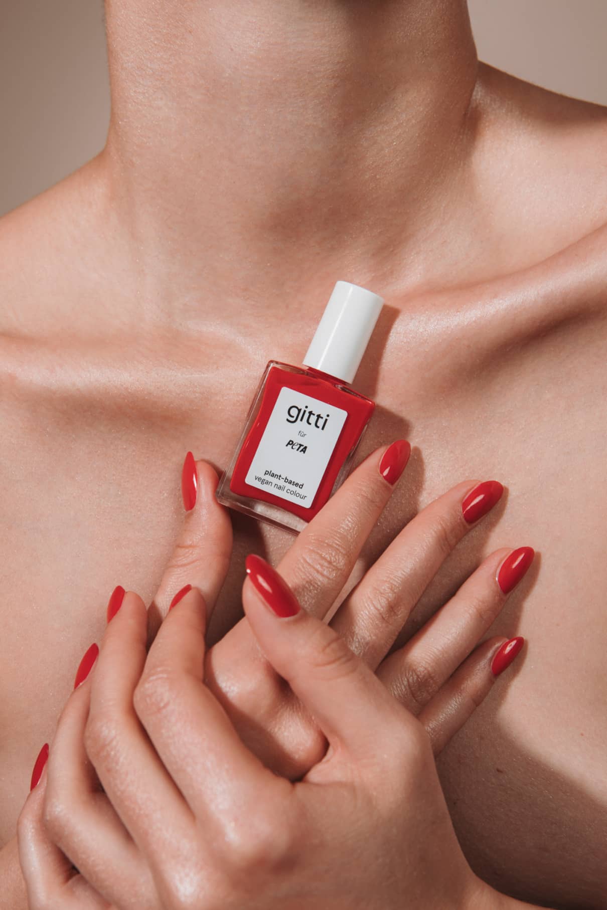 Eine Person mit rot lackierten Fingernägeln hält eine Flasche „gitti x PETA“-Nagellack in leuchtendem Rot mit orangefarbenen Untertönen vor ihre nackte Brust und ihr Schlüsselbein.
