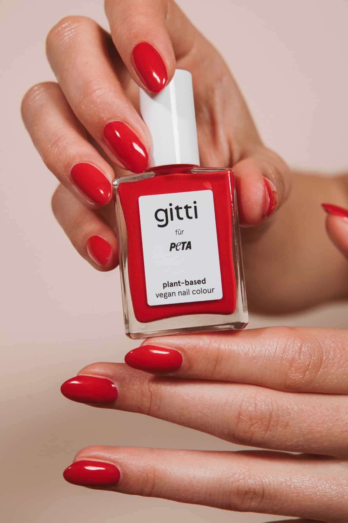 Eine Hand mit rot lackierten Nägeln in leuchtendem Rot mit orangefarbenen Untertönen hält eine Flasche „gitti x PETA“ – einen pflanzlichen, veganen Nagellack; darunter befindet sich eine weitere Hand mit passend lackierten Nägeln.