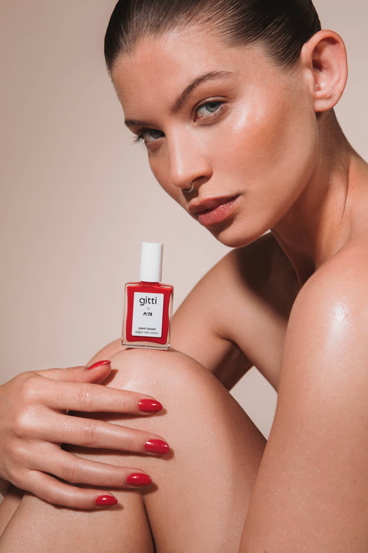 Eine Frau mit nach hinten geglätteten Haaren hält eine Flasche „gitti x PETA“-Nagellack in leuchtendem Rot mit orangefarbenen Untertönen in der Hand, der zu ihren manikürten Nägeln passt, während sie ihren Arm auf ihrem Knie ablegt – vor einem neutralen Hintergrund.