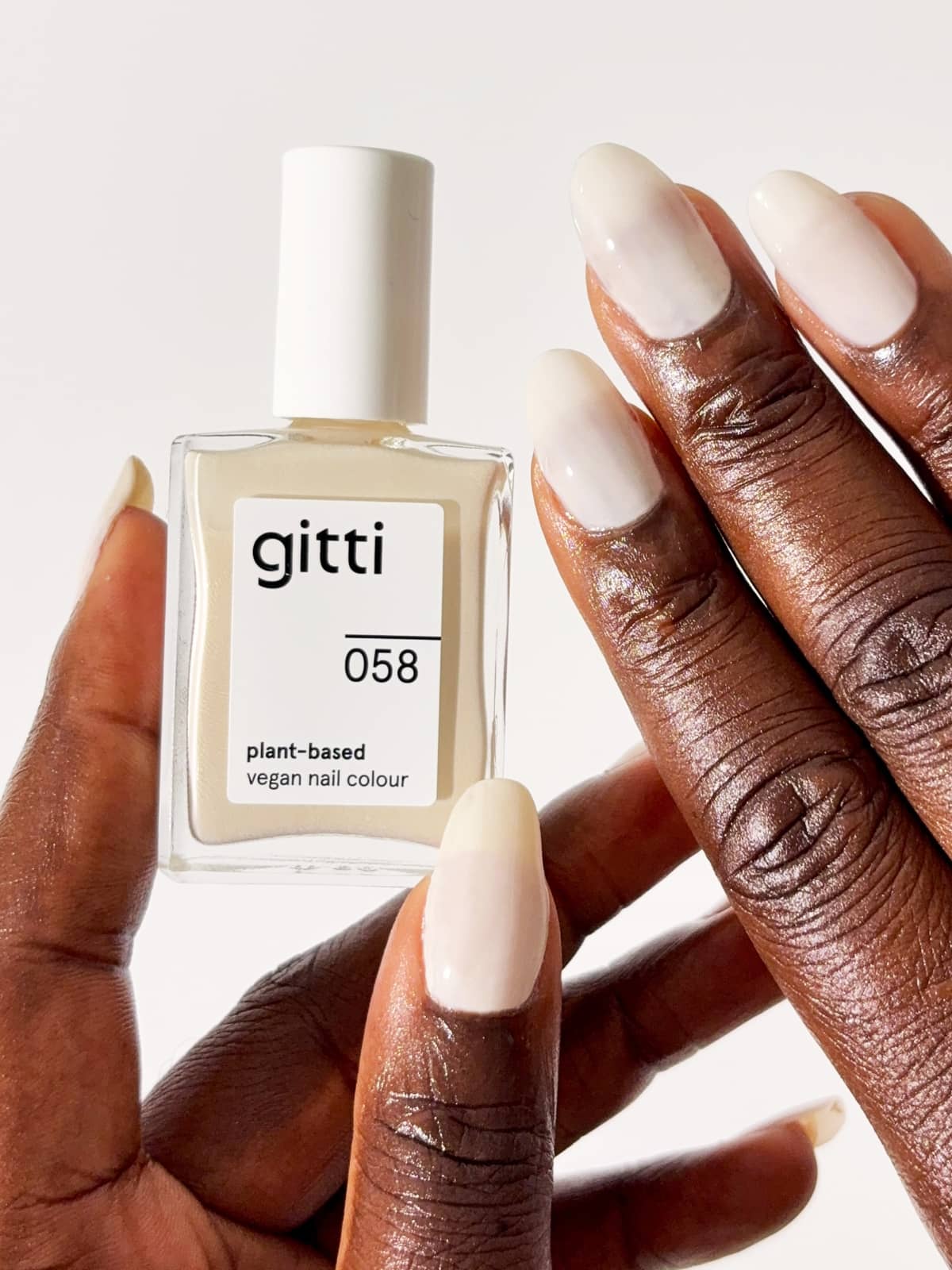 Eine Hand, die eine Flasche des pflanzlichen, veganen Nagellacks „Gitti Nr. 058“ hält, mit Nägeln, die in einem wärmeren Farbton lackiert sind – einem sandfarbenen, cremefarbenen Ton, der einen eleganten Look erzeugt und perfekt zu einem leichten, natürlichen Stil passt.