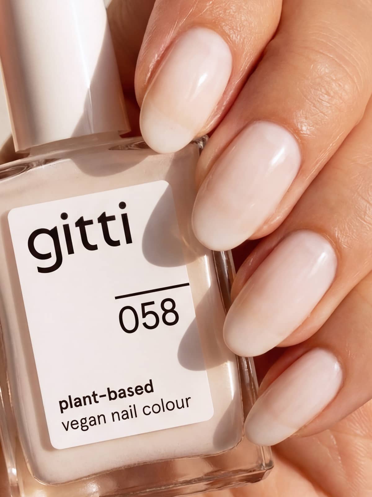 Eine Hand mit natürlich geformten Nägeln, lackiert in einem zarten Milchweiß aus dem „Colour & Concentrate Trio“, hält eine Flasche mit der Aufschrift „gitti 058 pflanzenbasierte Farben plant-based vegan nail colour“ in der Hand und präsentiert eine frische Maniküre, die durch das pflegende „Aloe Drop Nail Concentrate“ perfekt ergänzt wird.