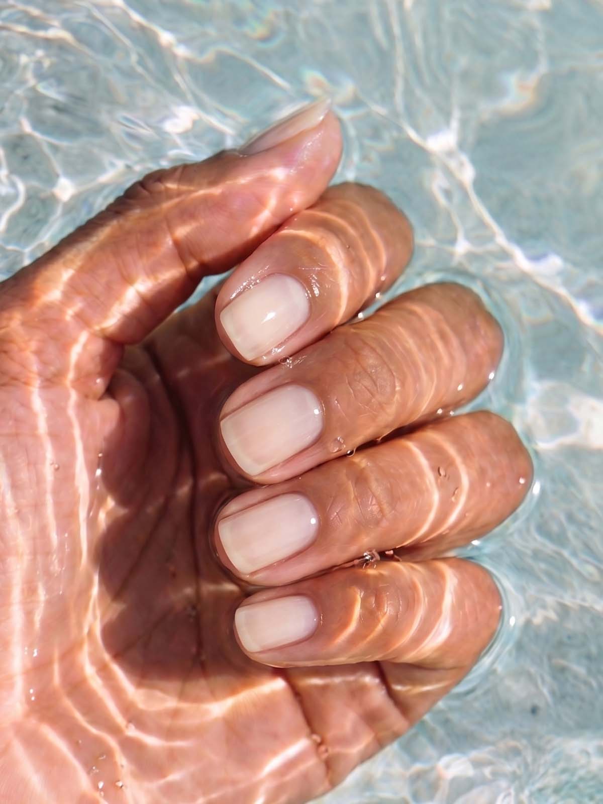 Eine Hand mit kurzen, gepflegten Naturnägeln taucht in klares Wasser ein, wobei das Sonnenlicht wellenförmige Reflexe auf der Haut und den Nägeln erzeugt. Der dezente Glanz der Lacke aus dem All-in-One-Set kommt wunderschön zur Geltung, unterstrichen durch eine makellose Grundierung, die jeden Nagel glatt und strahlend erscheinen lässt.