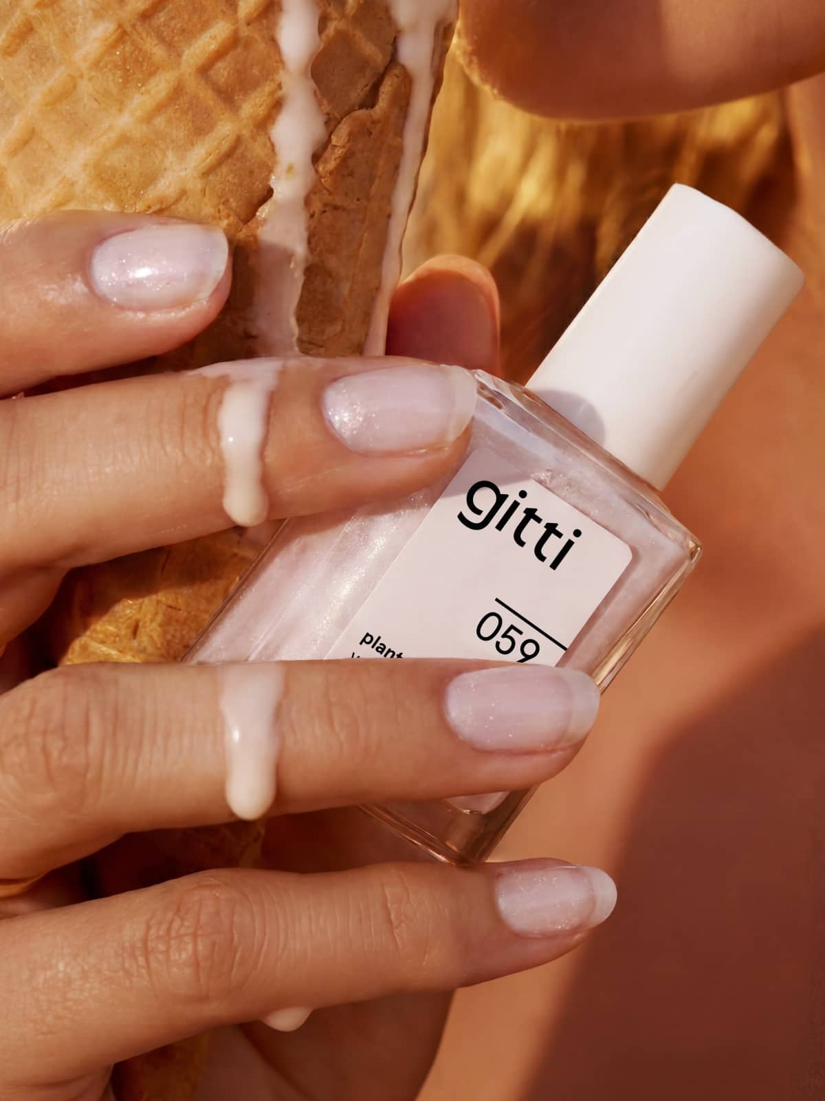 Eine Hand mit hellrosa, pflanzlichem Nagellack hält eine schmelzende Eistüte und eine Flasche „gitti Colour & Concentrate Trio“ mit der Aufschrift „059“.