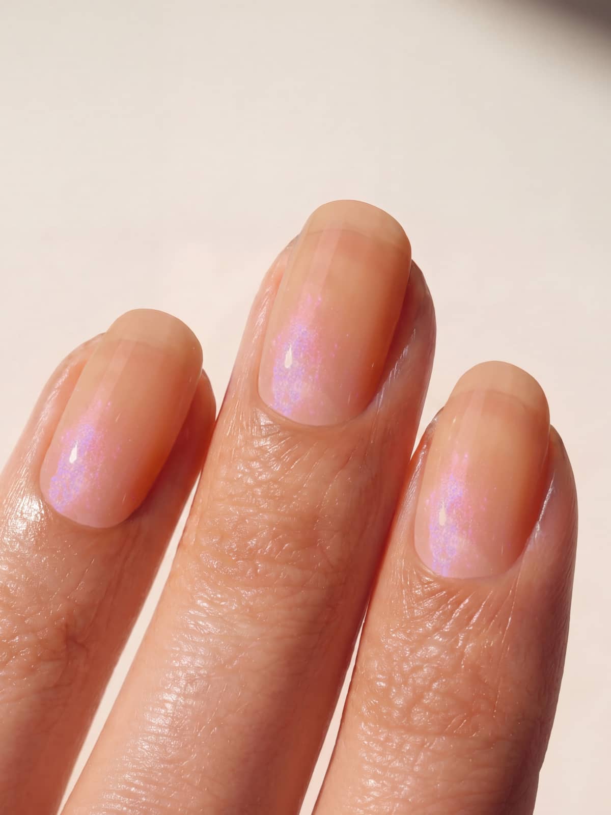 Eine Nahaufnahme von drei Fingern mit sorgfältig gepflegten Nägeln, lackiert mit „Gitti Nr. 060“ – einem transparenten, glänzenden rosa Nagellack, der das Licht mit einem kühlen Schimmer einfängt, während sein transparenter apricotfarbener Farbton den Nägeln einen frischen Glanz verleiht.
